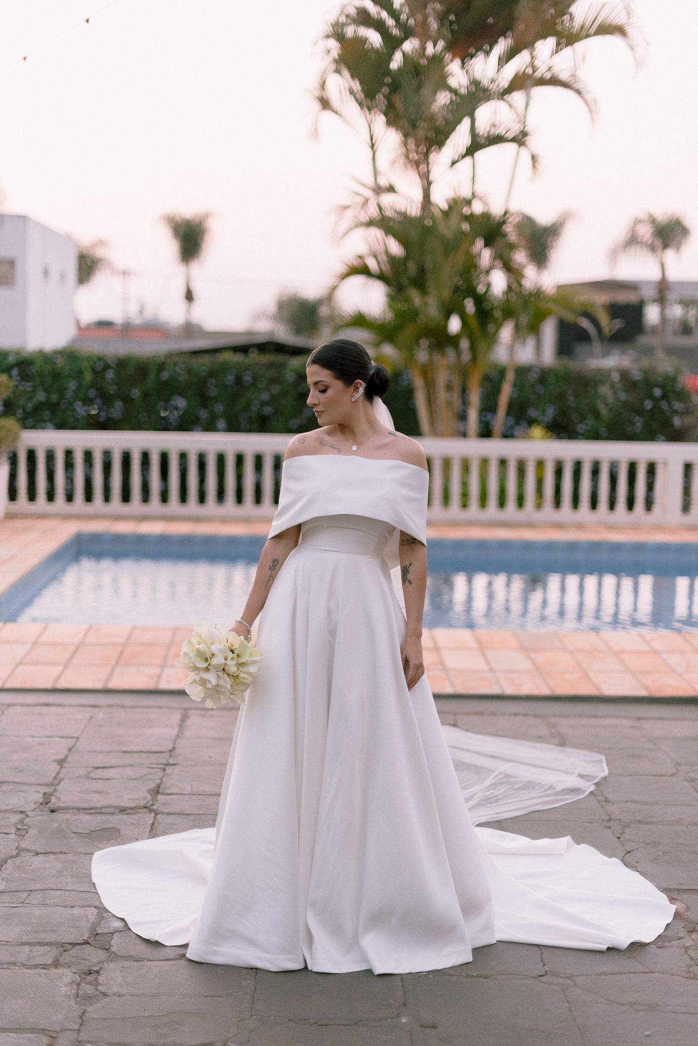 Bride in an elegant off-shoulder ballgown poses by a swimming pool during sunset.