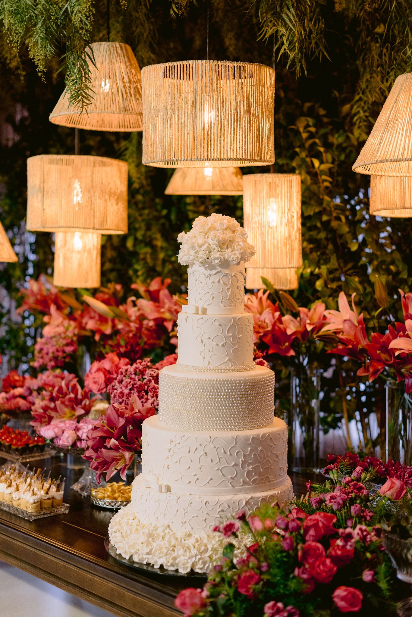 An elegant white wedding cake with floral details surrounded by hanging wicker lamps and pink flowers.