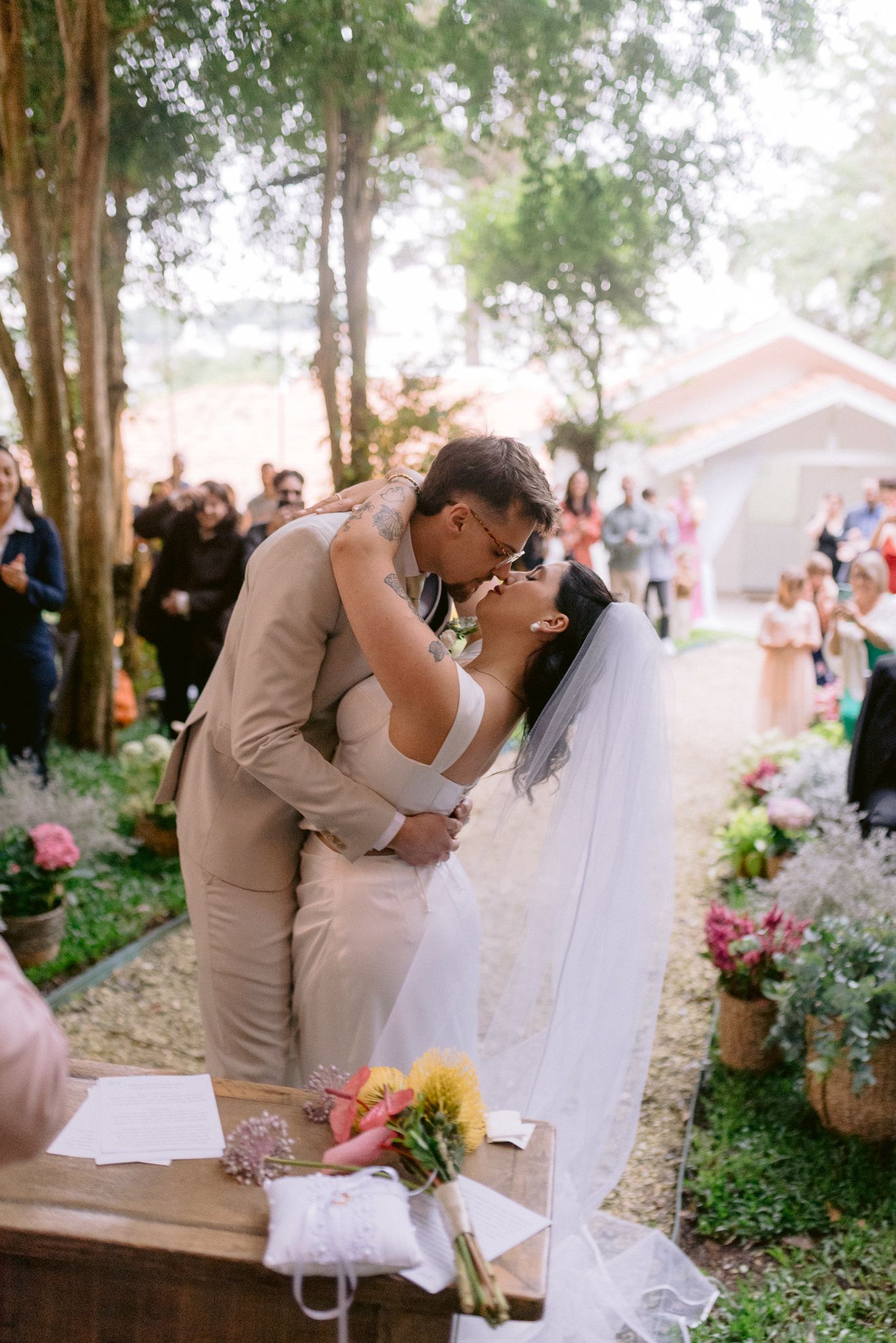 A joyful wedding couple shares romantic kisses in a garden setting with trees and pink flowers.