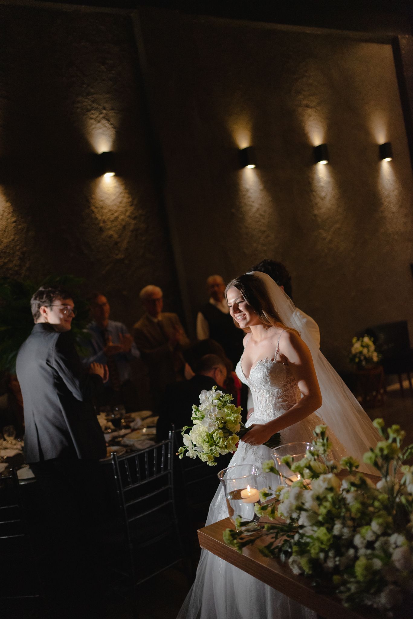 A wedding reception setup in a dimly lit venue with elegant floral arrangements and mood lighting along the walls.
