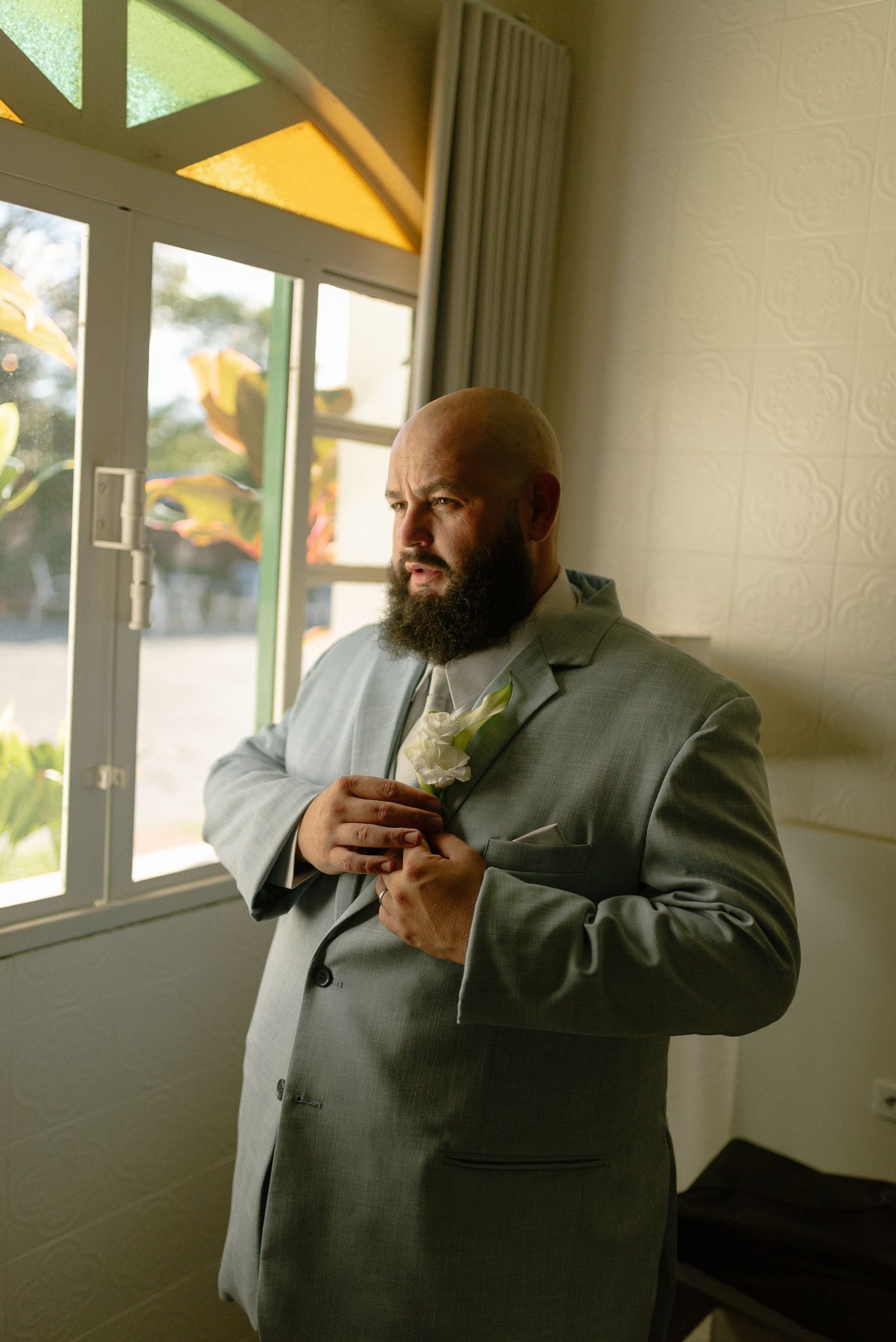 A bearded man in a formal gray suit adjusts his tie while looking out a window.