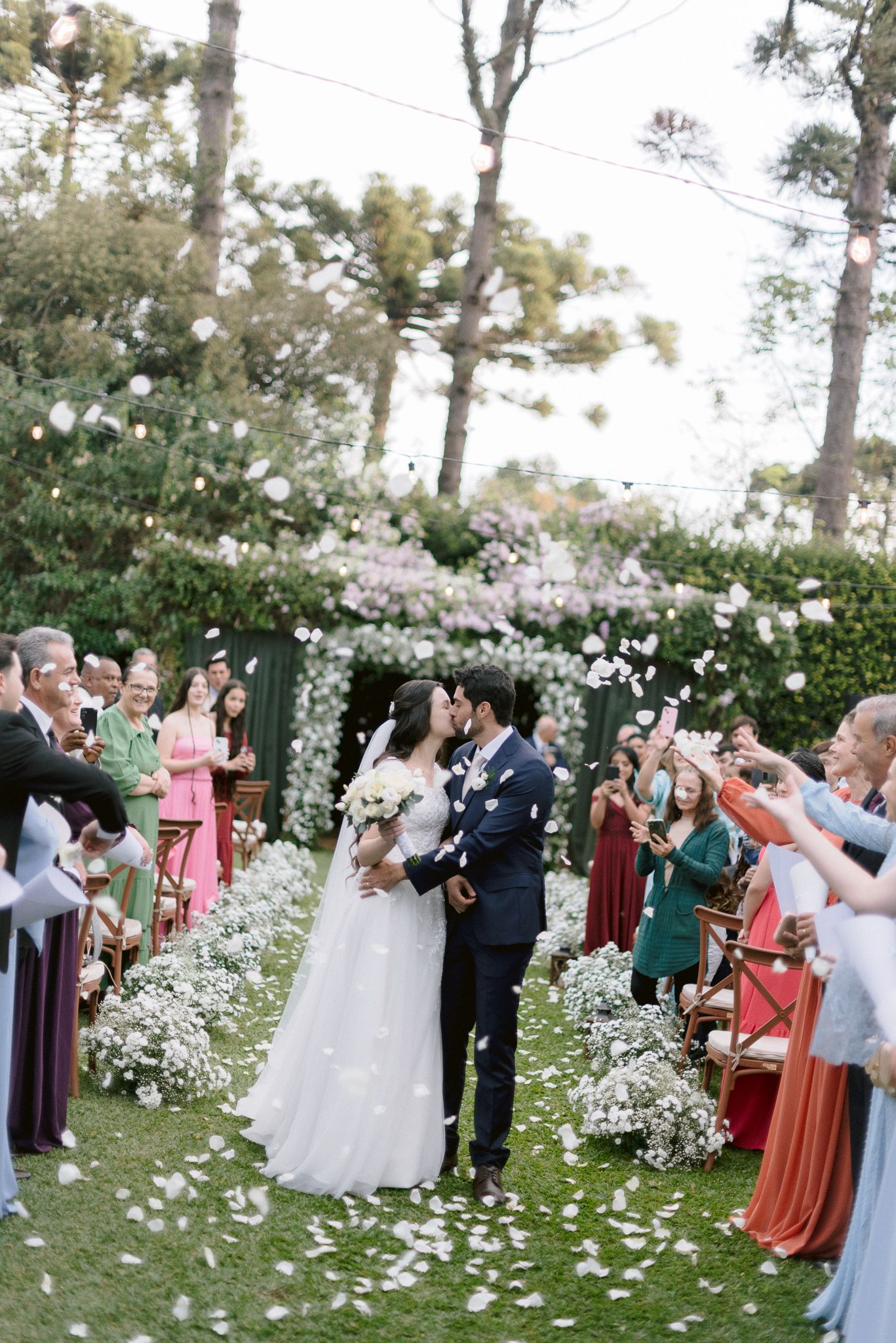 Wedding guests celebrate in an outdoor garden ceremony surrounded by white flowers and tall pine trees.