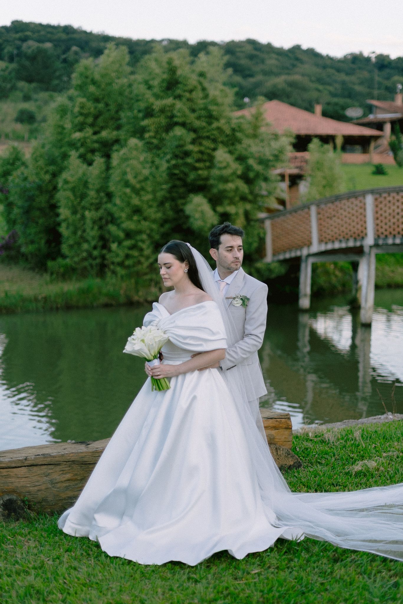 Couple sharing romantic moments by a serene lake against lush greenery and rustic buildings in the background.