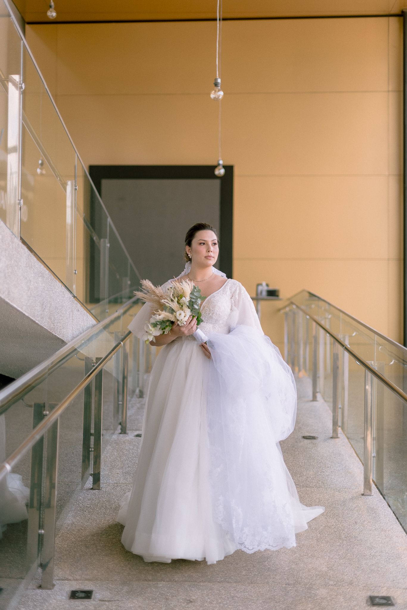 A bride in a flowing white wedding gown gracefully poses along a modern glass and metal staircase.