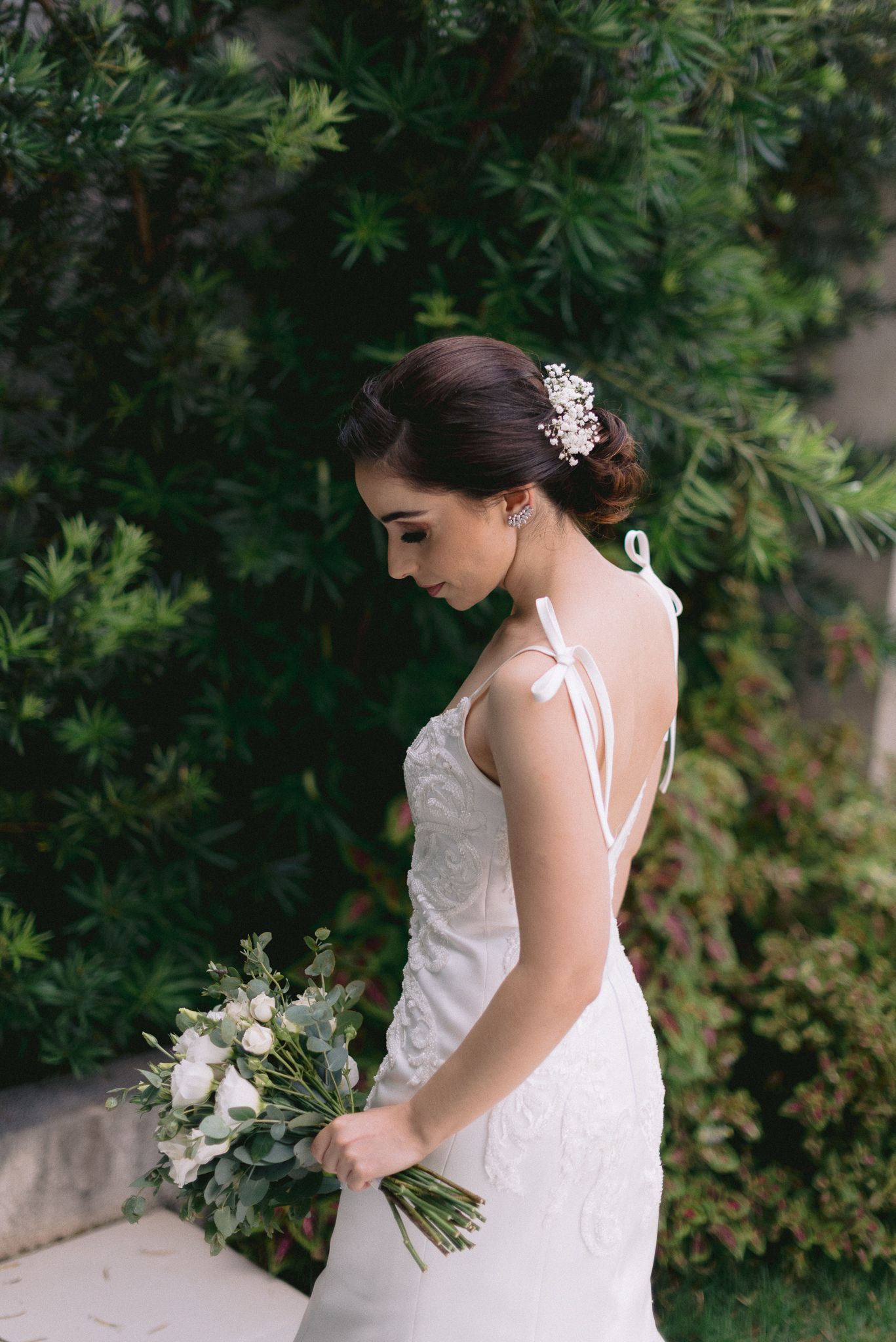Side profile of bride in white dress with ribbon straps against greenery.