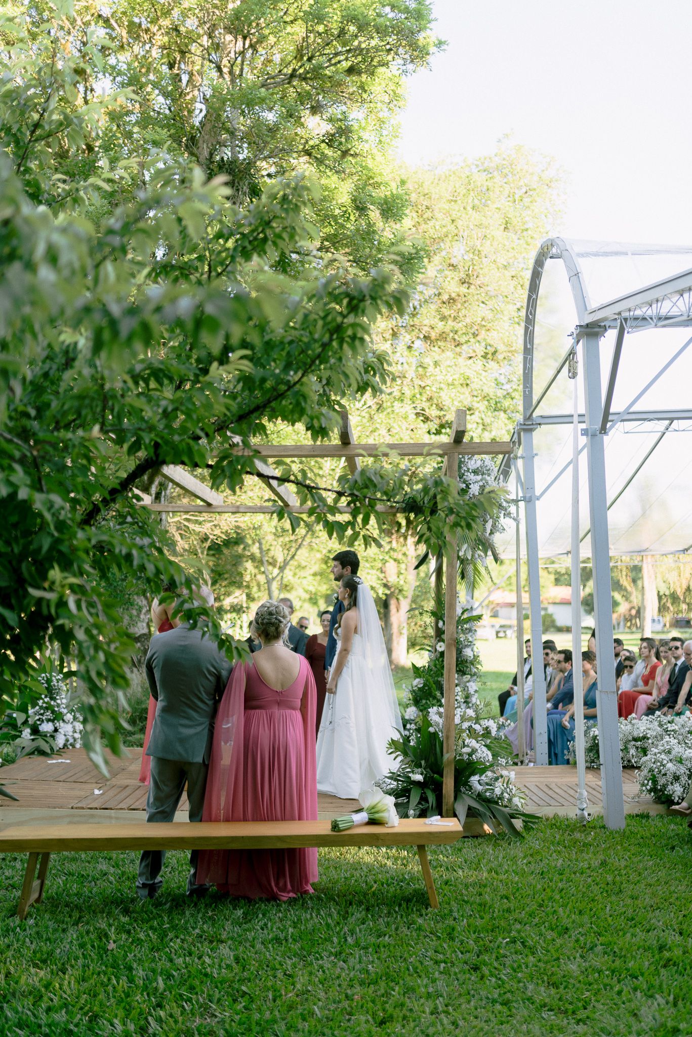 Wedding guests gather under a lush green arbor for an outdoor ceremony overlooking a scenic bridge.
