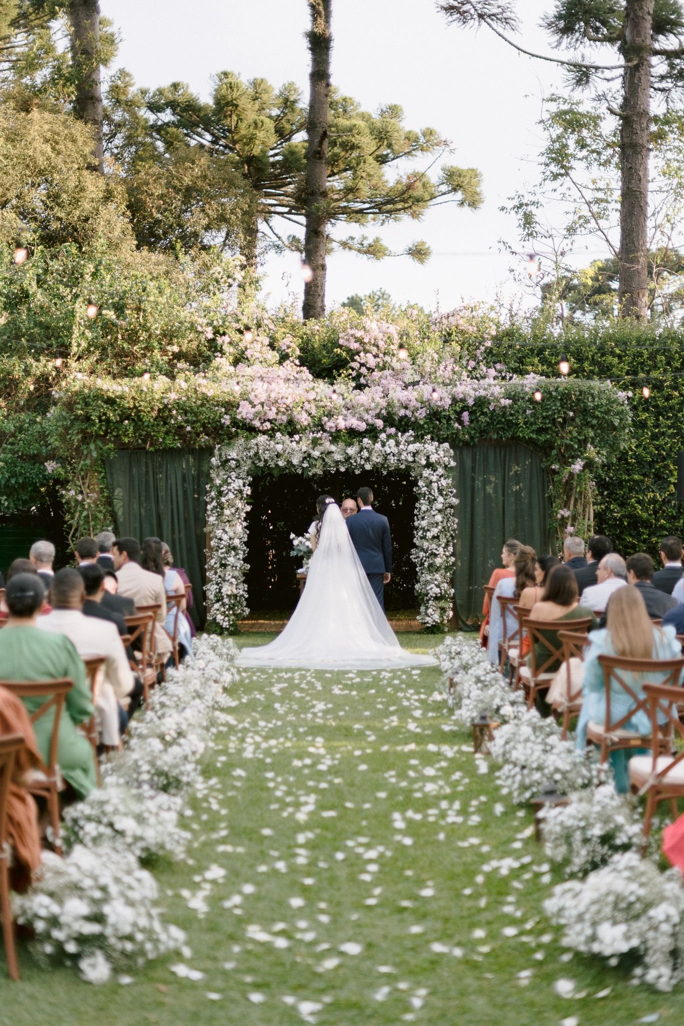 Outdoor wedding ceremony setting with white floral aisle decorations and guests seated on a lush lawn.
