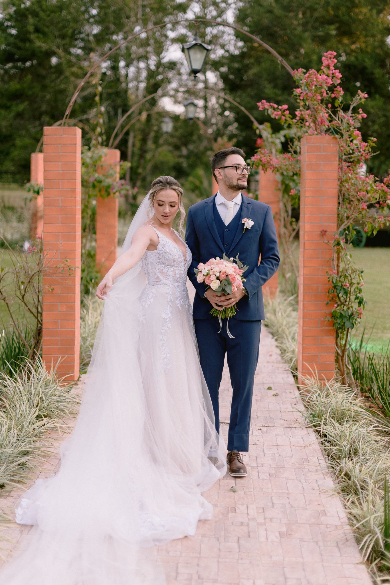 Couple walks along brick-lined garden path with coral flower bouquet.