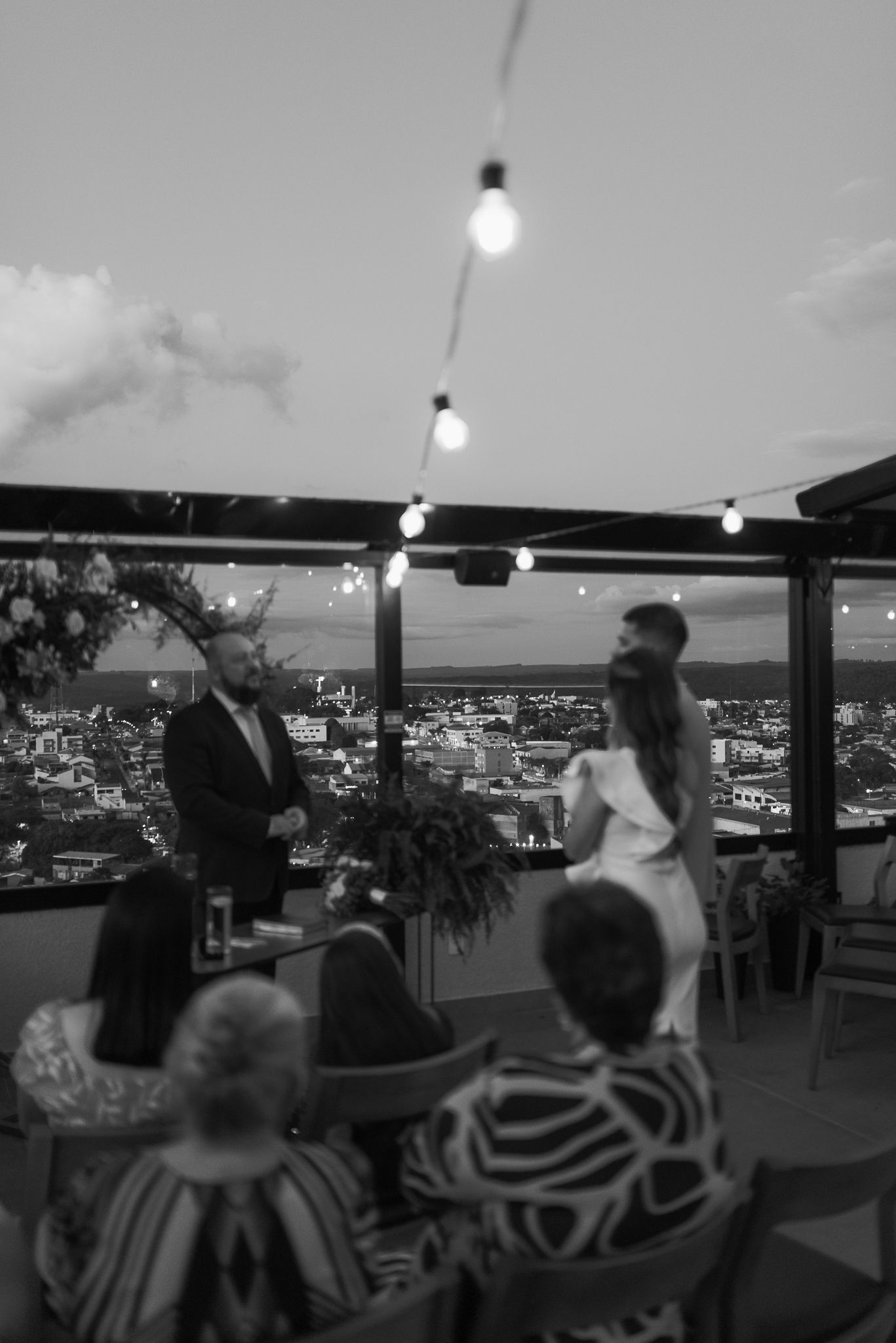 People dancing at a rooftop venue with city lights glowing in the background and string lights overhead.