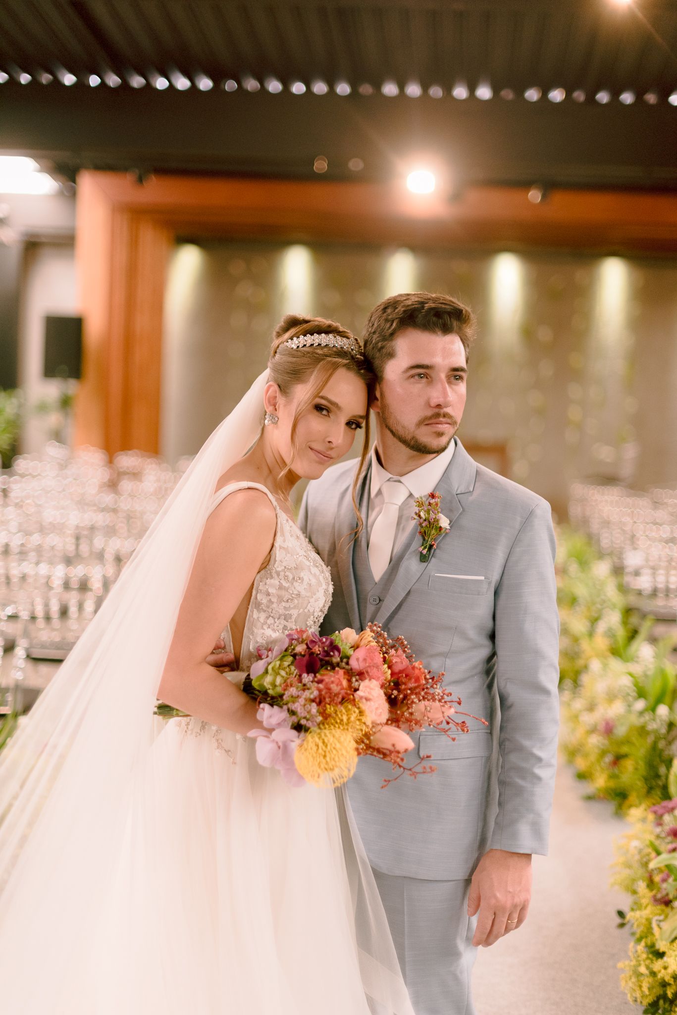 Wedding couple posing together in formal attire with colorful autumn bouquet against warm lighting.