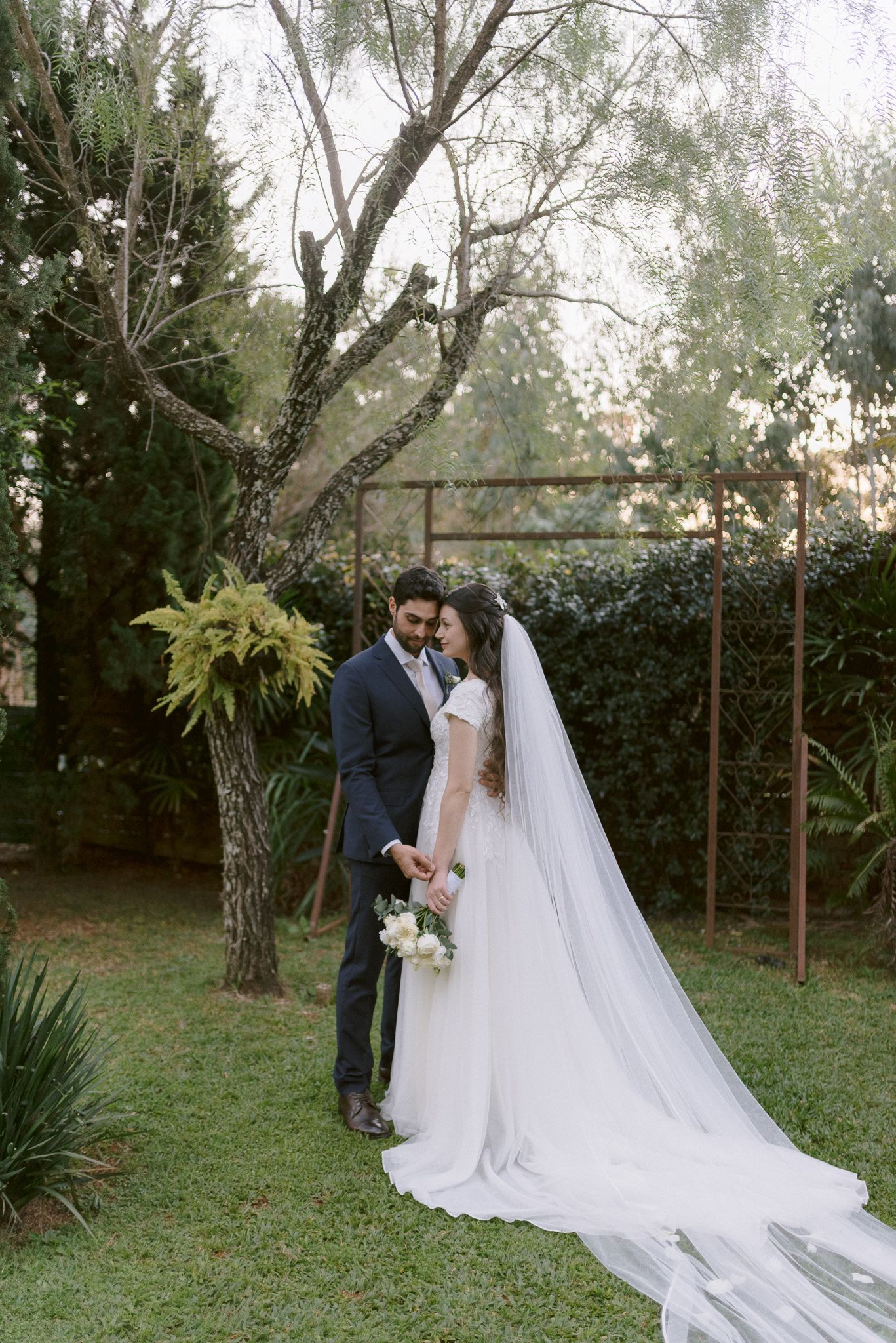 A romantic outdoor garden wedding scene with a flowing white veil trailing behind a couple among tall trees.