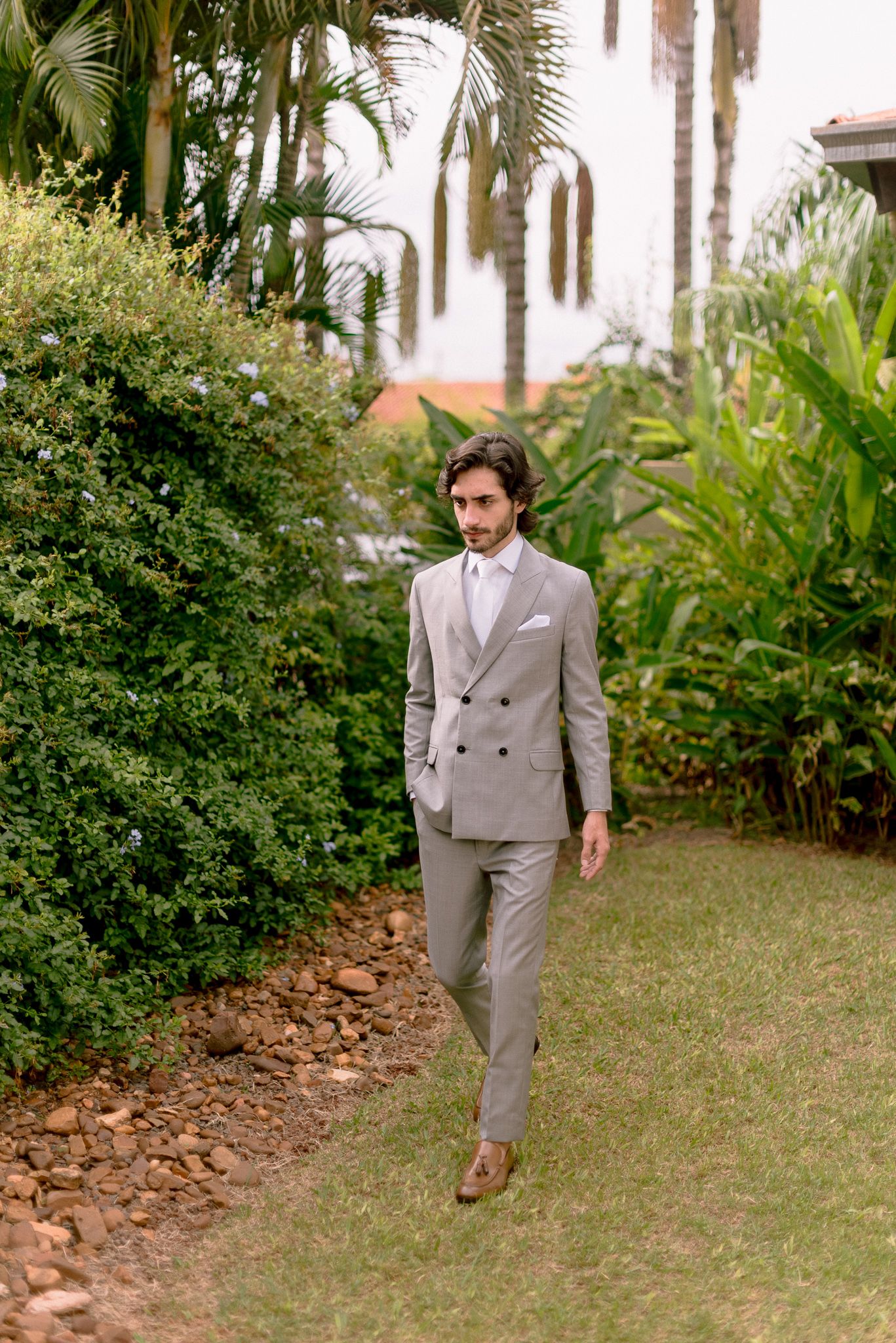 A person in a light gray suit walks along a garden path lined with tropical palm trees and lush greenery.