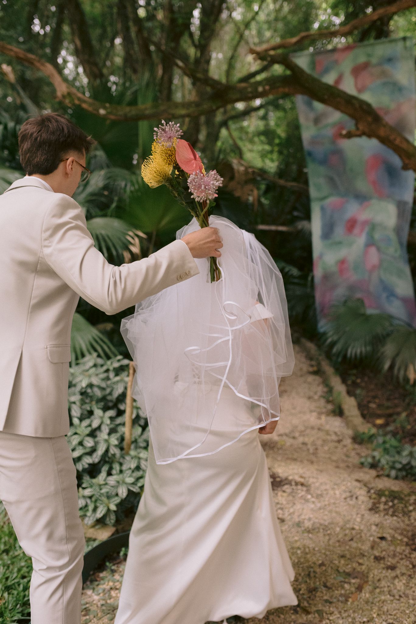 Wedding couple walks through lush garden setting in all-white attire.