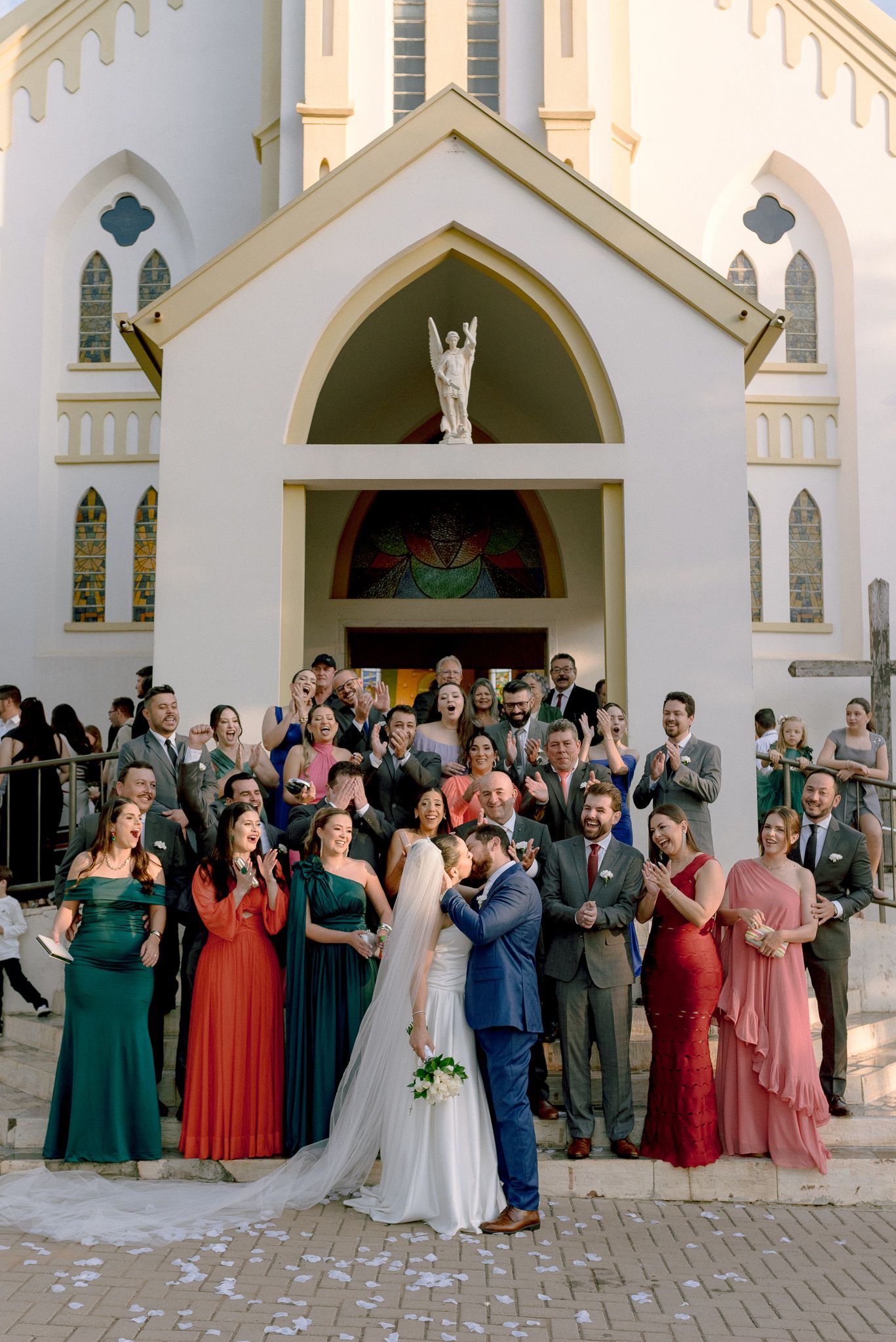Wedding party and guests celebrate with raised arms outside a Gothic style chapel with white arched doorways.