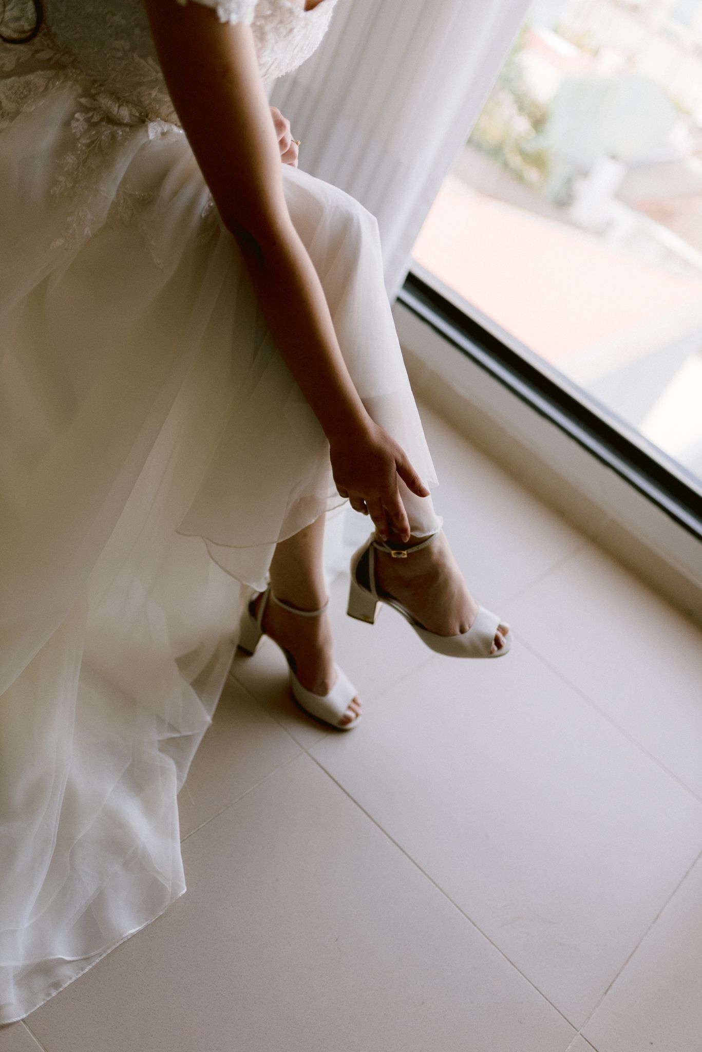 White wedding dress hem and shoes seen in a delicate backlit detail shot against a light flooring.