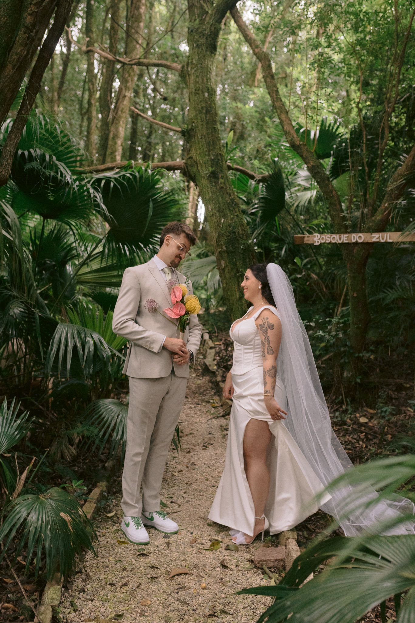 Wedding couple poses playfully in a tropical forest setting with palm fronds and natural light.
