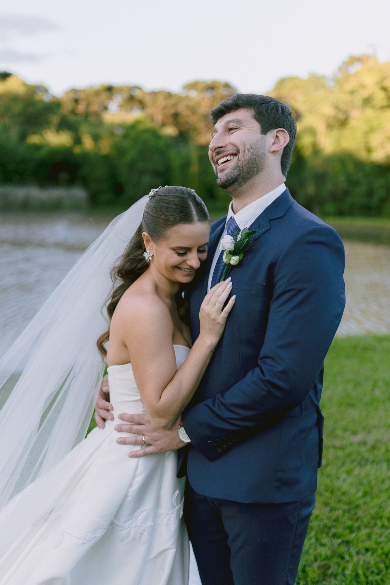 A joyful wedding couple embraces outdoors with a flowing white veil and navy suit against a natural backdrop.