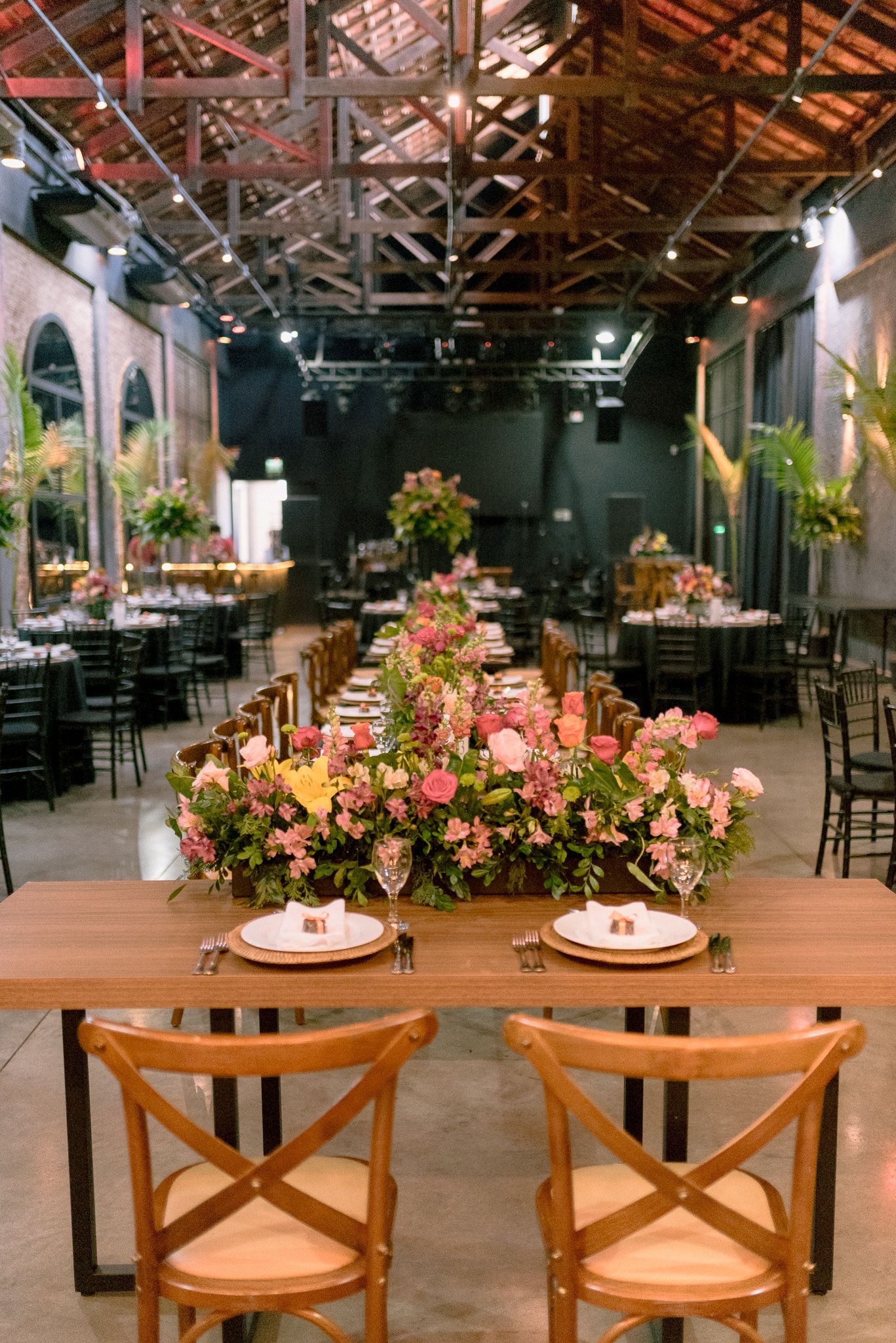 Elegant wedding reception venue with exposed wooden beams, black chairs and pink floral centerpieces on dining tables.