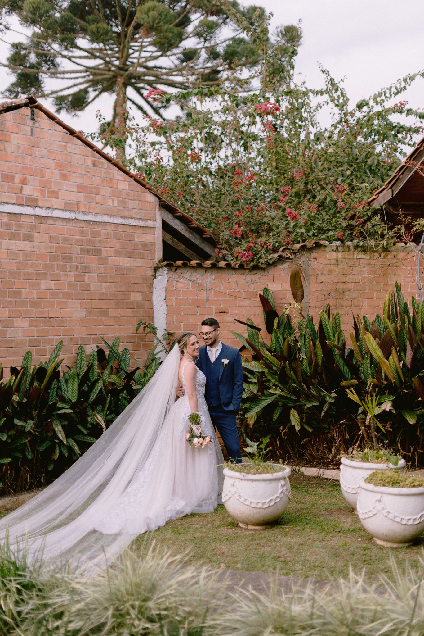 A wedding couple poses in an elegant garden setting with araucaria trees and decorative planters.