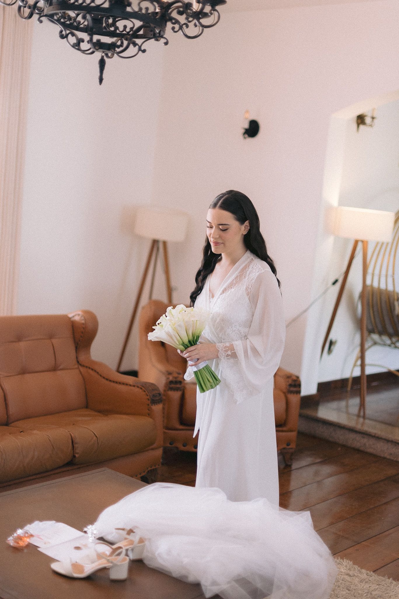 A bride in white silk robe relaxes in an elegant living room setting with brown furniture and soft lighting.