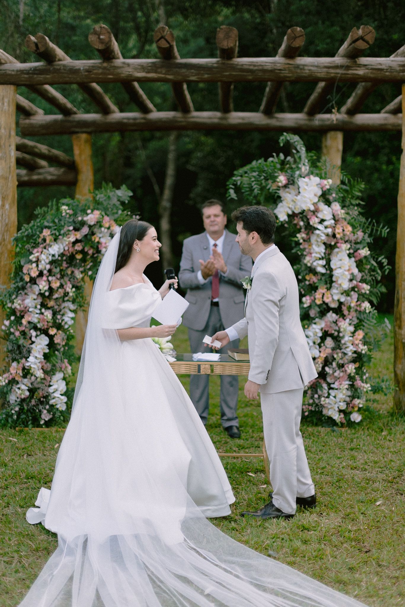 A romantic outdoor wedding ceremony unfolds under a rustic wooden arch adorned with pink and white floral arrangements.