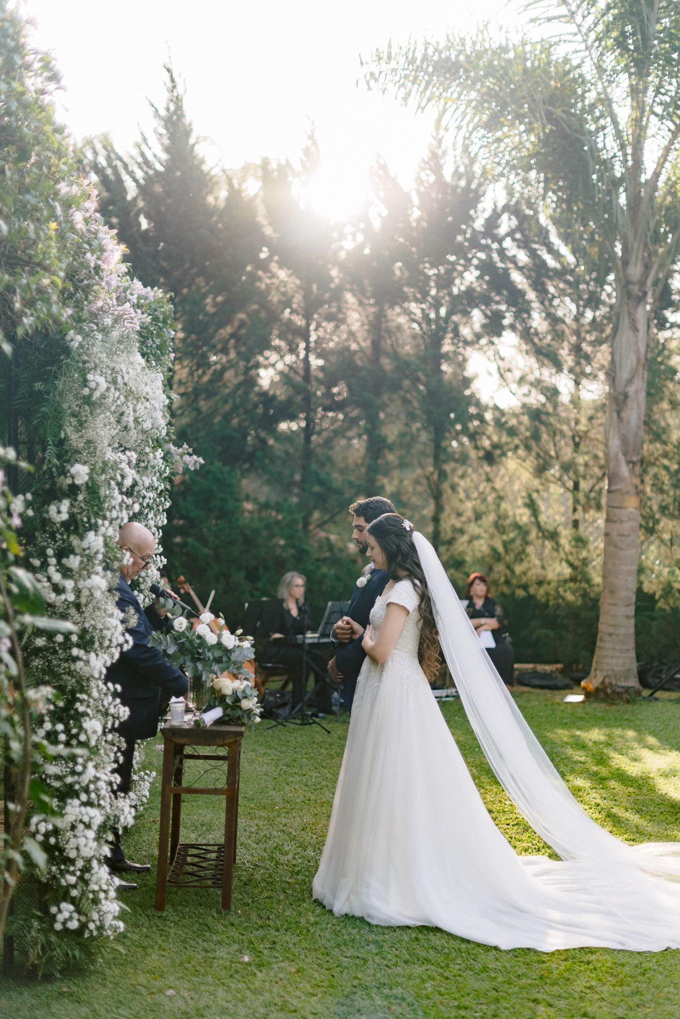 A bride in a flowing white gown walks towards a wooden archway decorated with white florals in a sunlit garden.