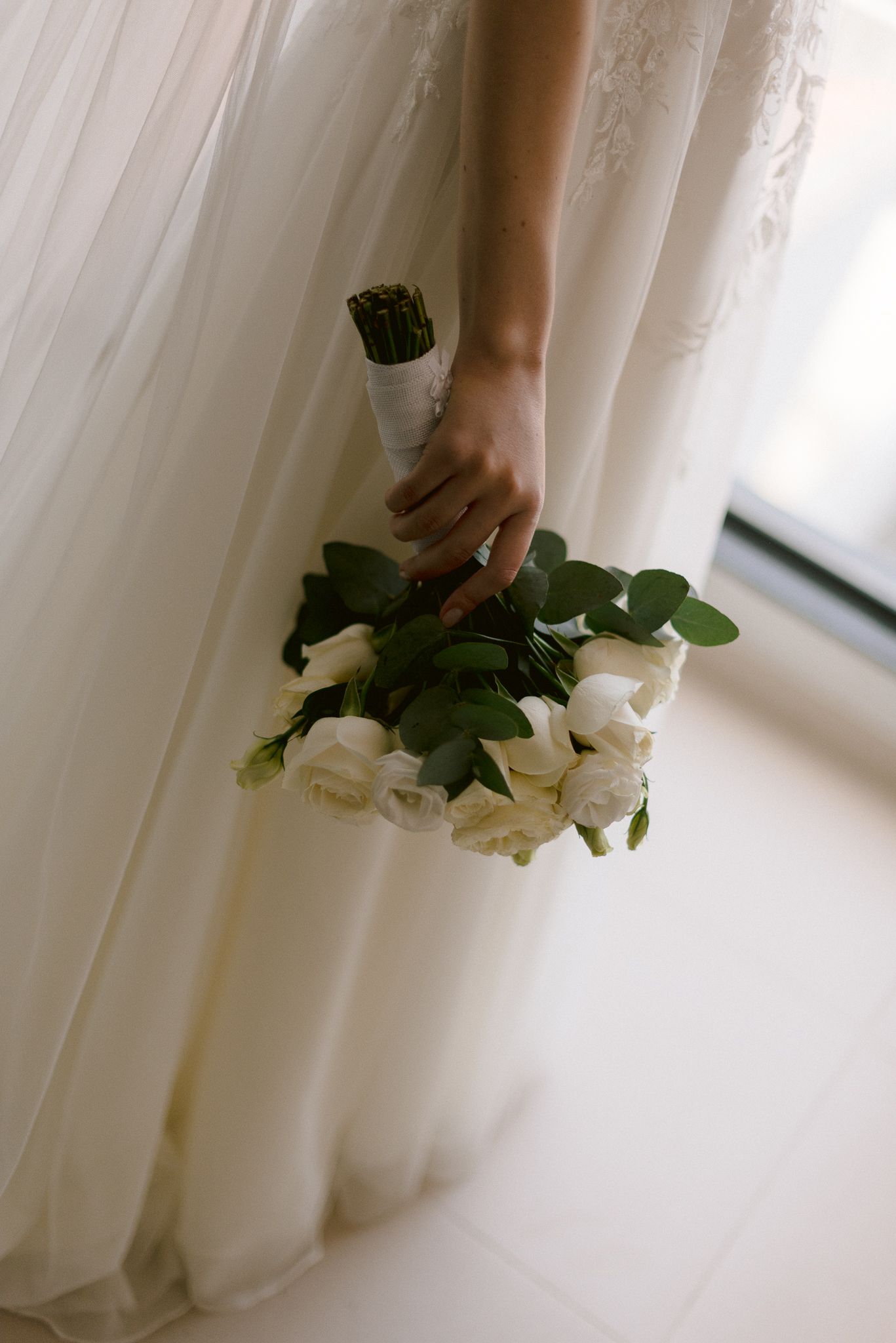 A close-up view of hands holding a delicate white rose bouquet against a soft, light curtain background.