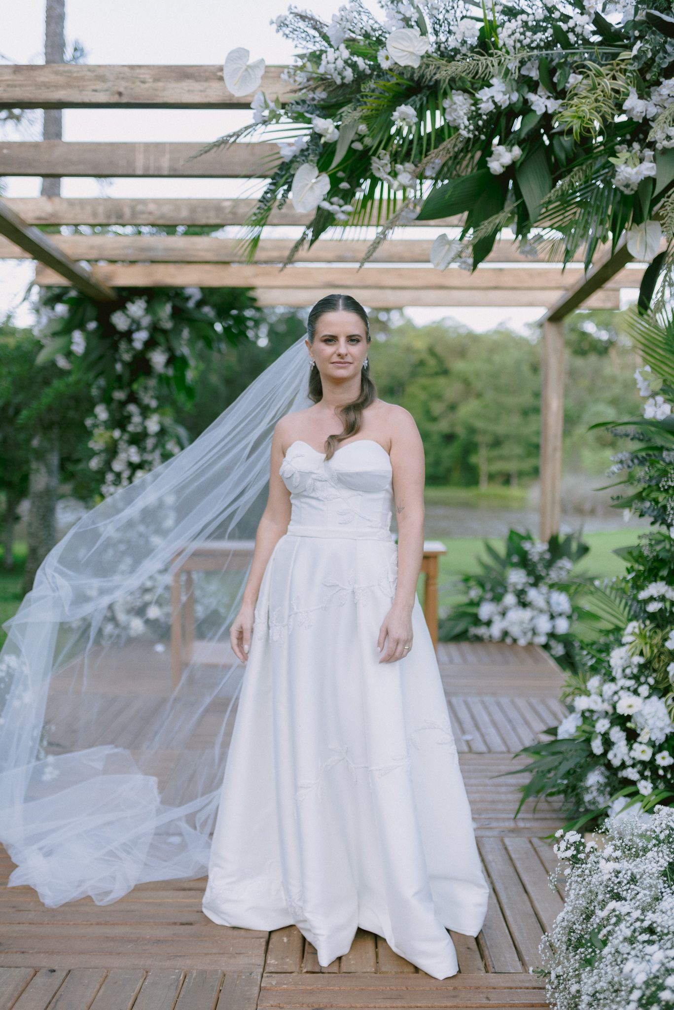 A bride in a white wedding dress stands on a wooden deck surrounded by delicate white floral arrangements and greenery.