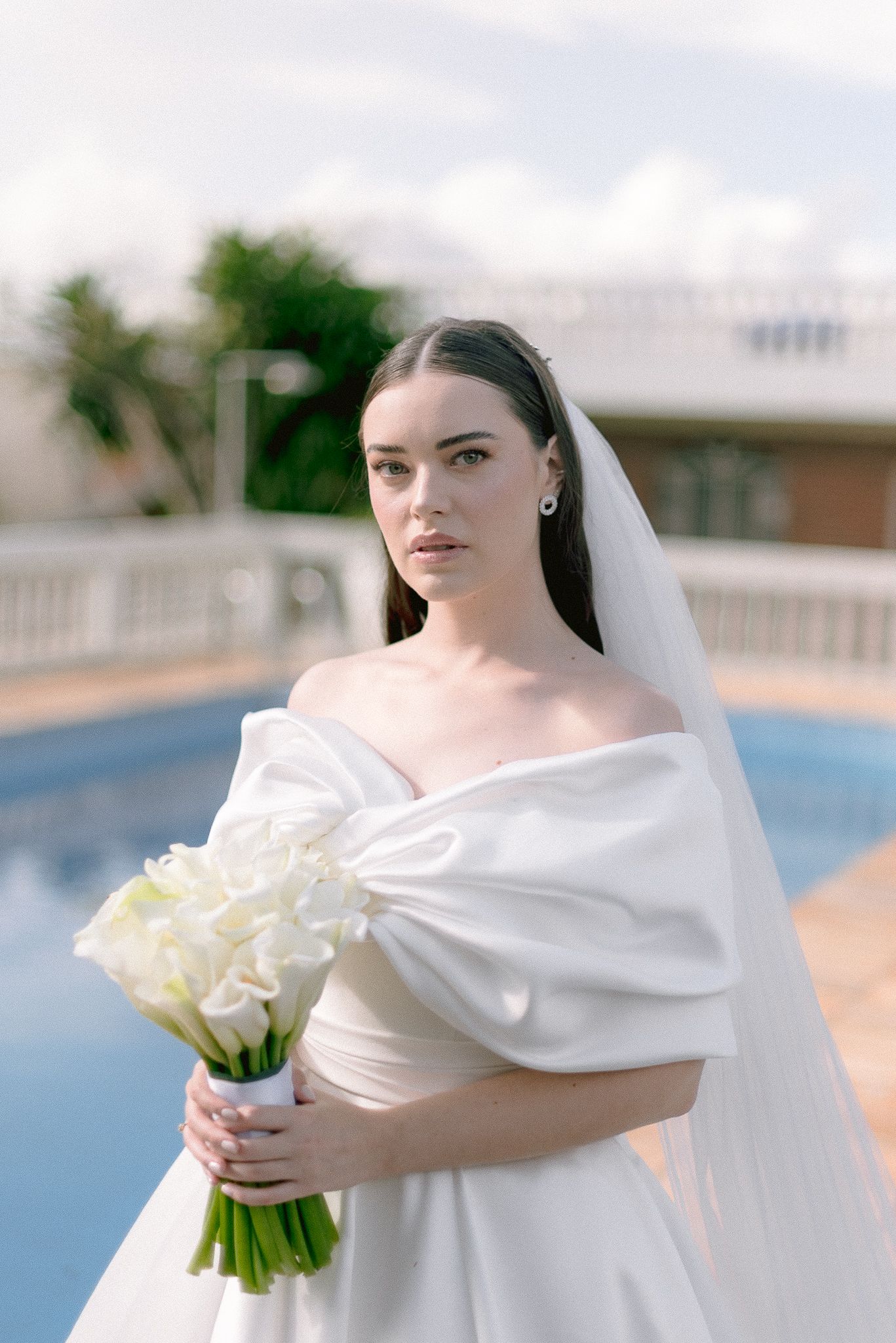 A bride in an elegant white off-shoulder wedding dress holds a bouquet of white calla lilies by a pool.