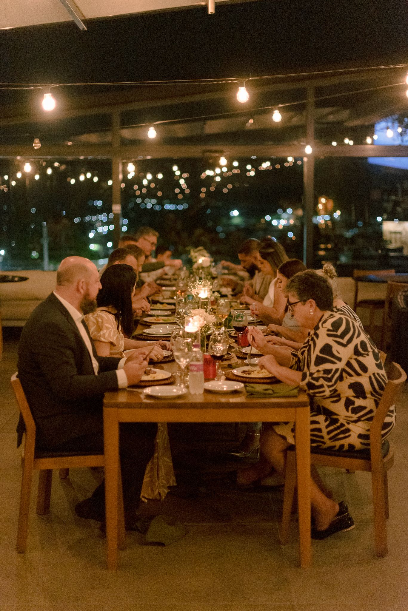 Diners enjoy an intimate candlelit dinner at a long wooden table under string lights with city lights twinkling in background.