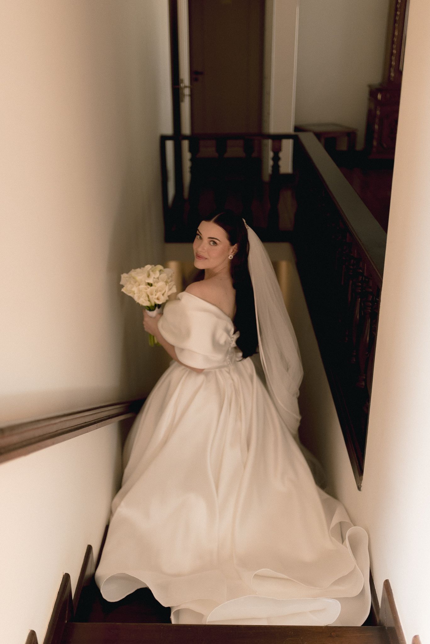 A bride in flowing white dress and veil sits gracefully in a minimalist hallway with natural light.
