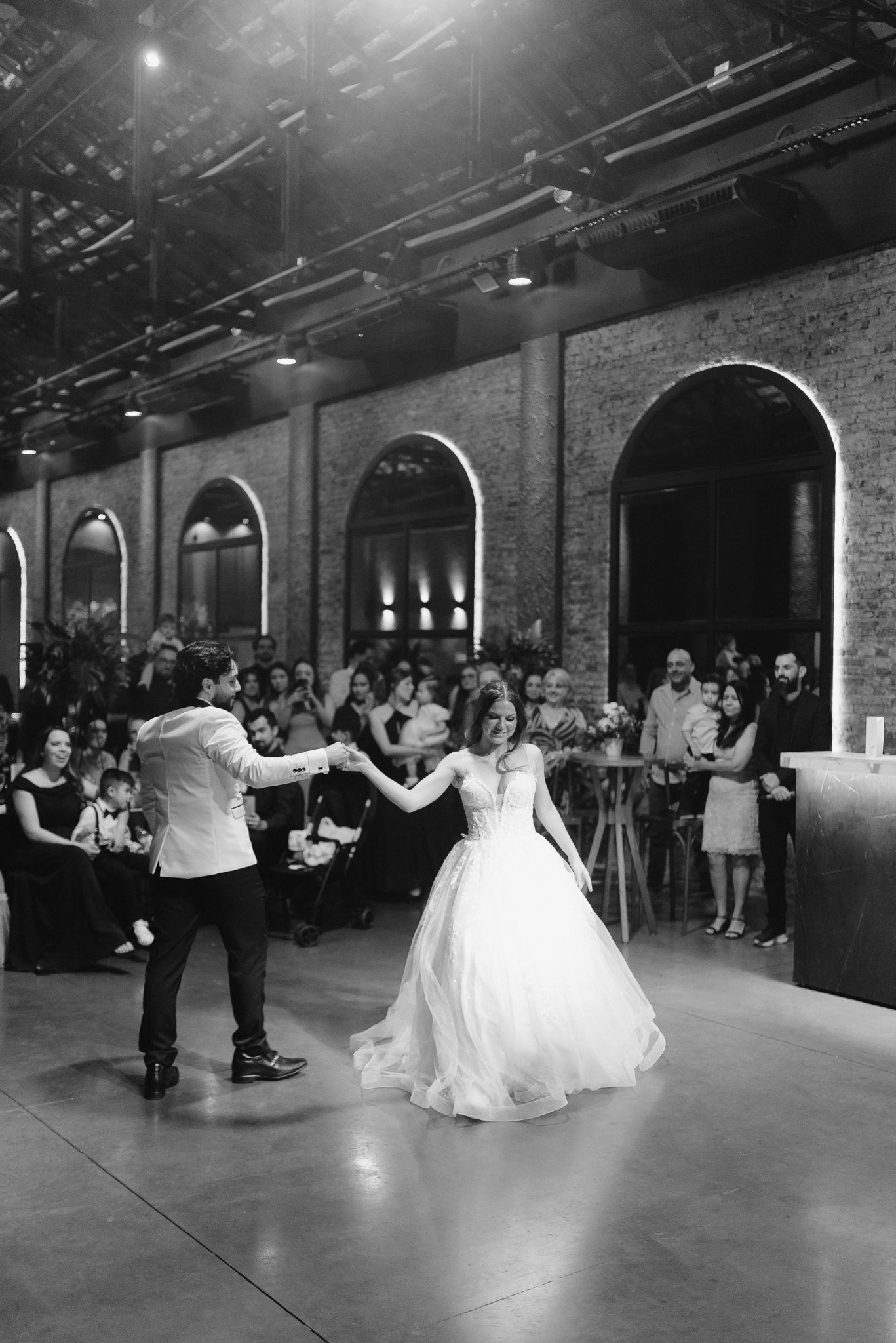 Couples dance together during a wedding reception in a spacious brick-walled venue with arched windows.