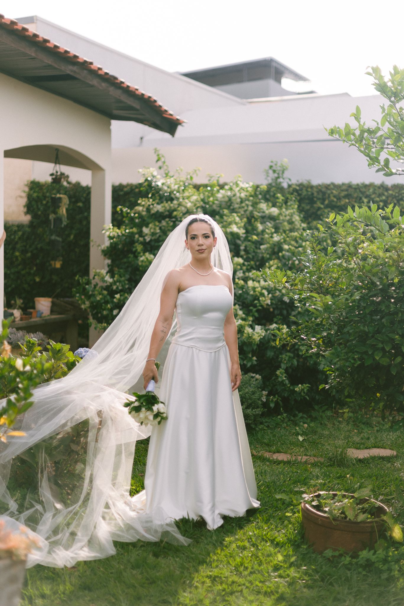 Bride in white empire waist gown and flowing veil poses in lush garden setting for bridal portraits.