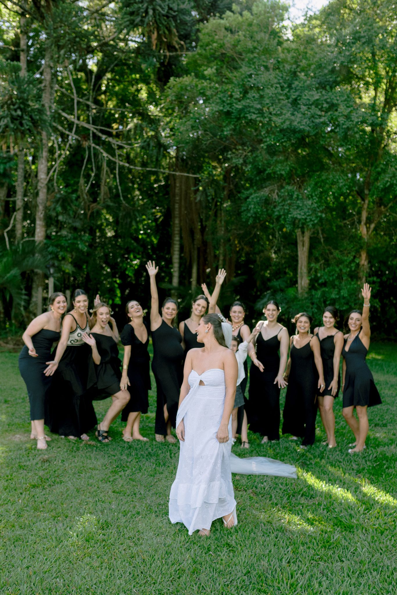 A group of bridesmaids in black dresses and a bride in white pose playfully in a wooded outdoor setting.