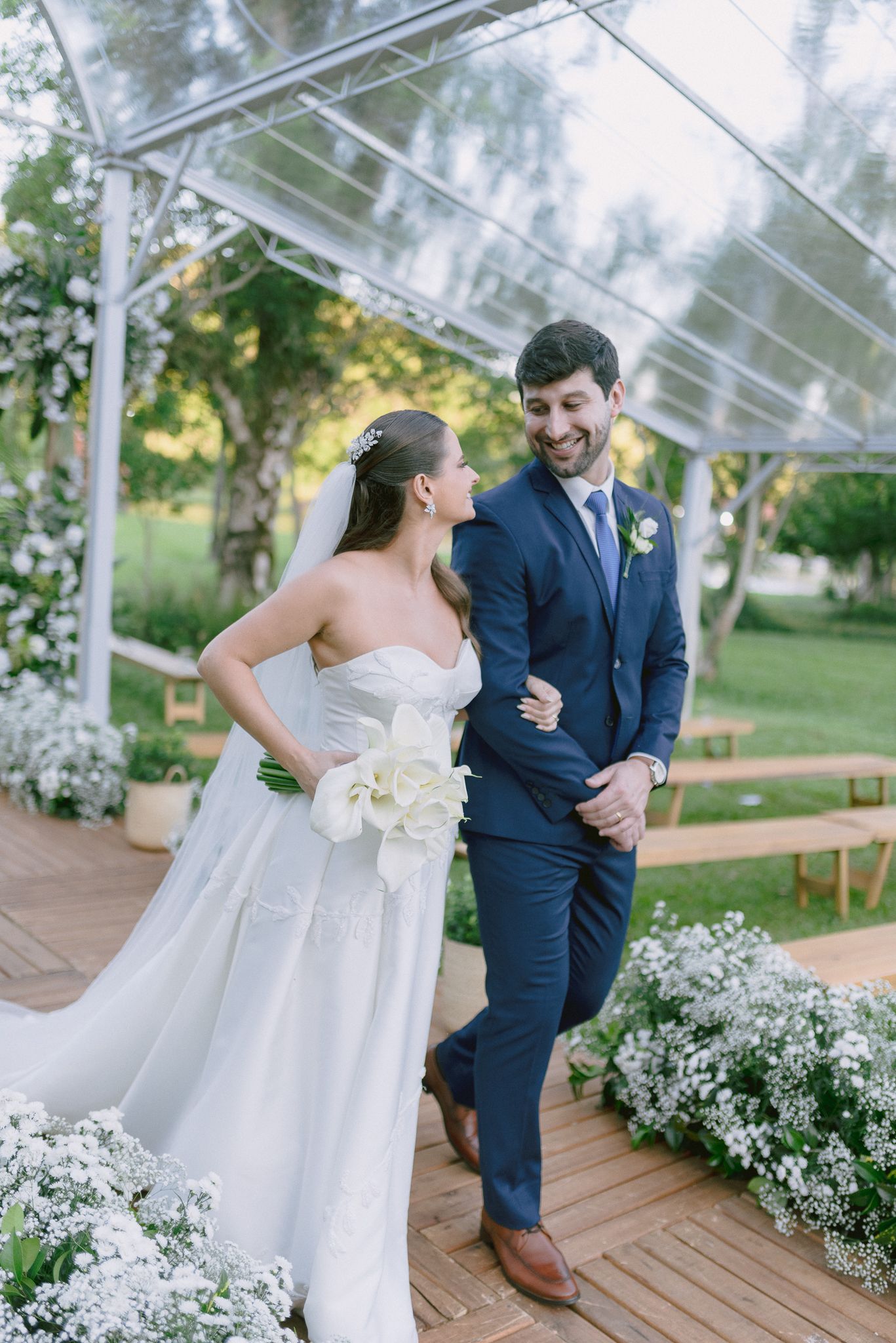 A couple shares a joyful moment on a wooden deck surrounded by white flowers and greenery at their outdoor wedding.