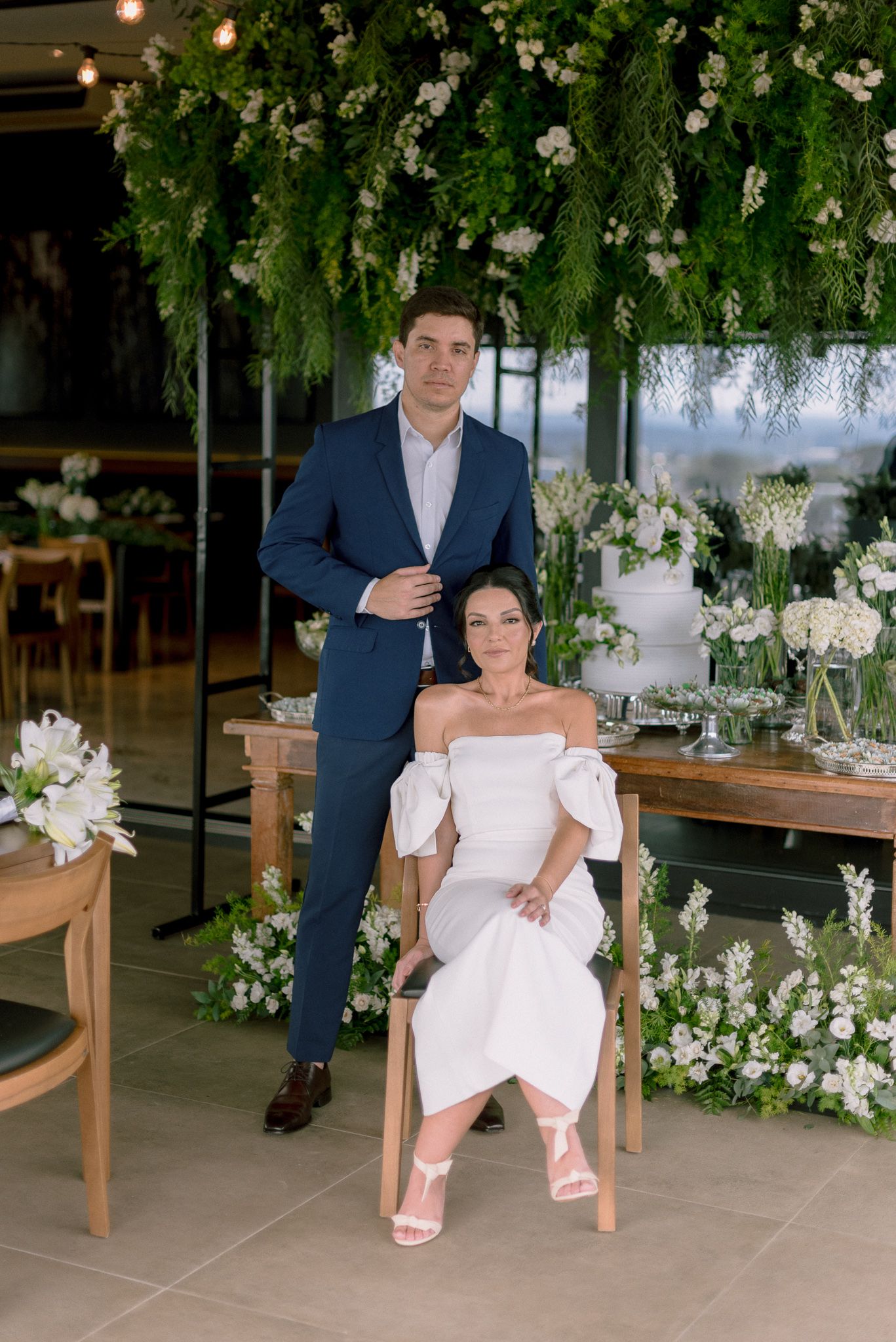 Wedding couple seated in wooden chairs under a decorative floral canopy.