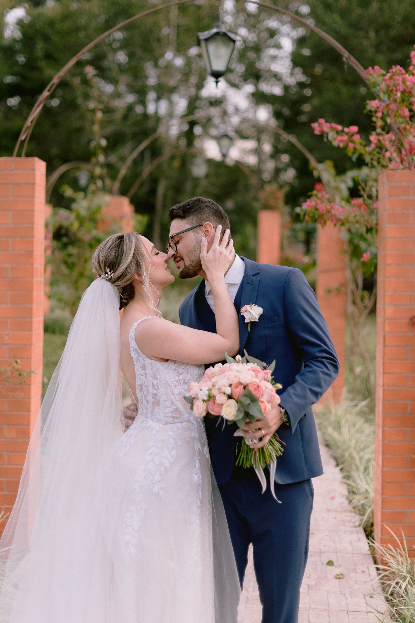 A romantic wedding couple shares a tender moment in a garden setting surrounded by brick pillars and pink flowers.