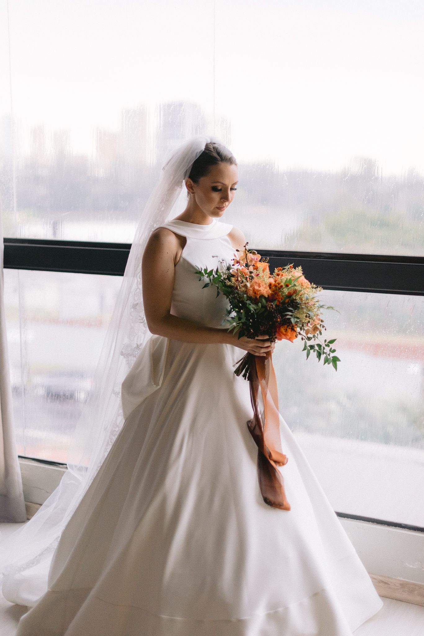 A bride in a white ballgown poses elegantly by large windows with a coral and peach floral bouquet.