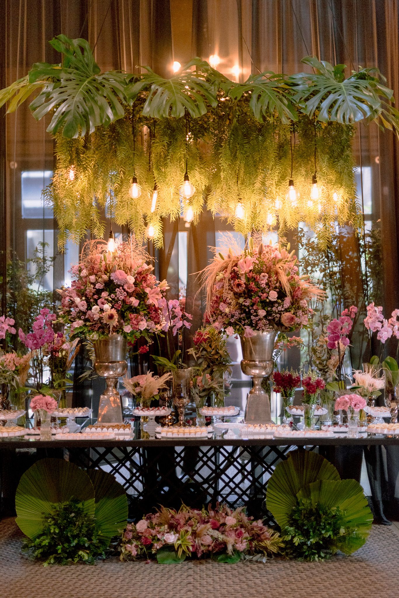 Elegant wedding reception decor features hanging greenery and pink floral arrangements under warm lighting.