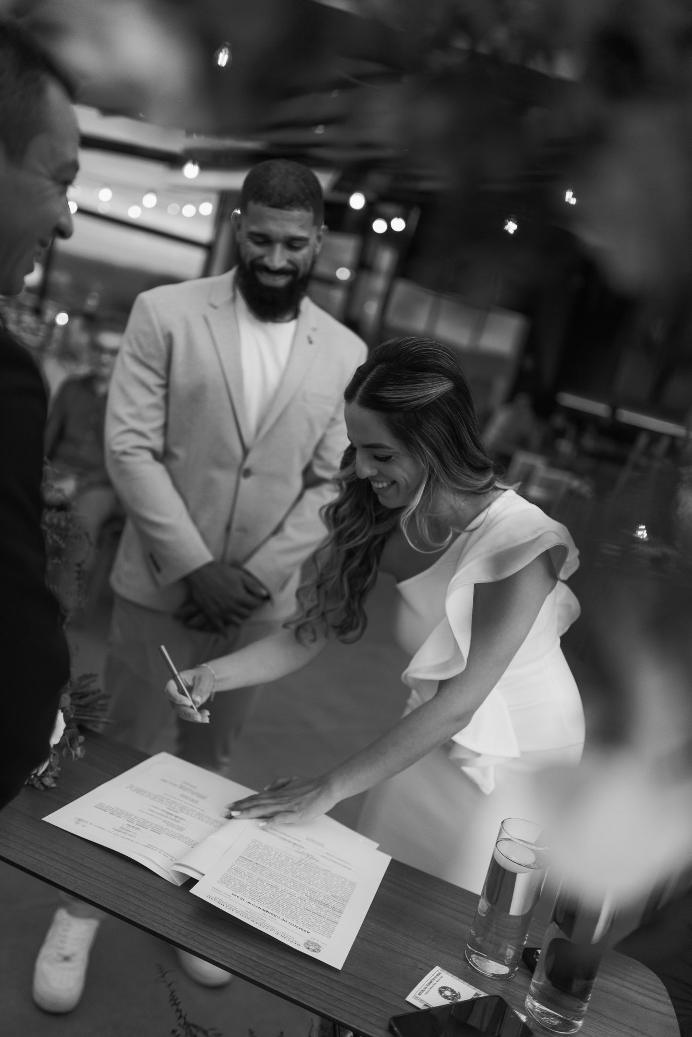 Black and white photo of a couple signing documents at a table during what appears to be a wedding ceremony.