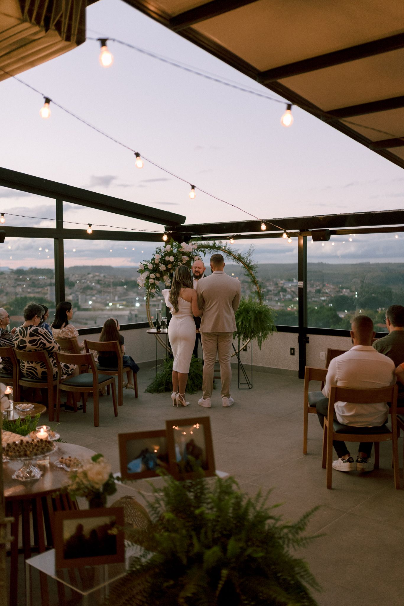 A covered rooftop venue decorated with string lights and greenery overlooking a scenic mountain landscape at dusk.