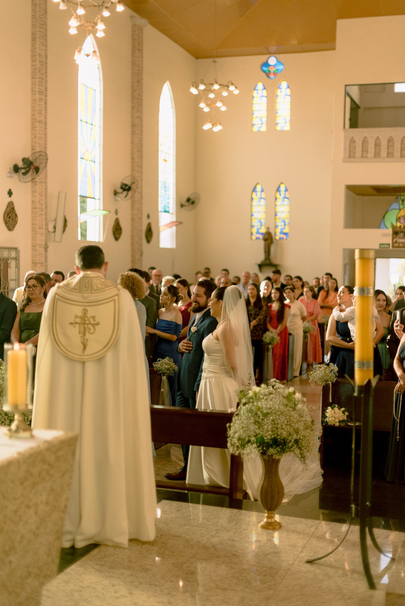 Wedding ceremony taking place in a bright church sanctuary with guests and floral arrangements along the aisle.