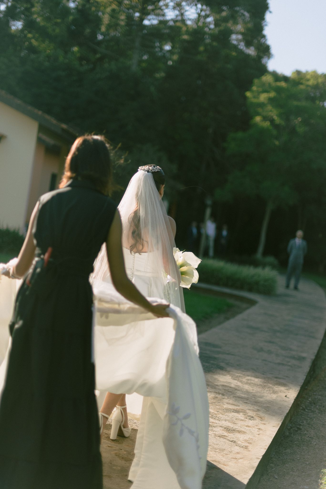 A bride in a white dress and veil walks alongside someone in black on a garden path during an outdoor wedding ceremony.