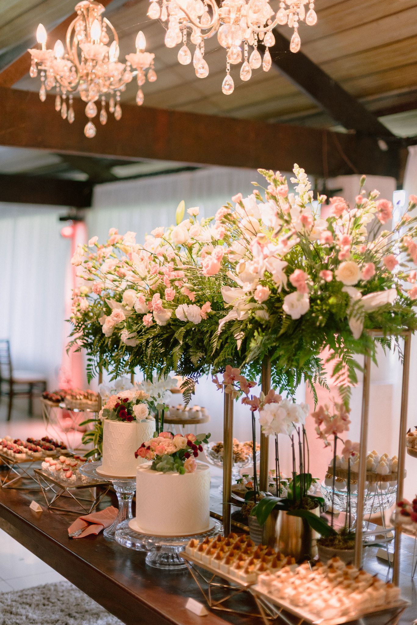 Luxurious wedding dessert table with white cakes, floral arrangements, and crystal chandeliers overhead.