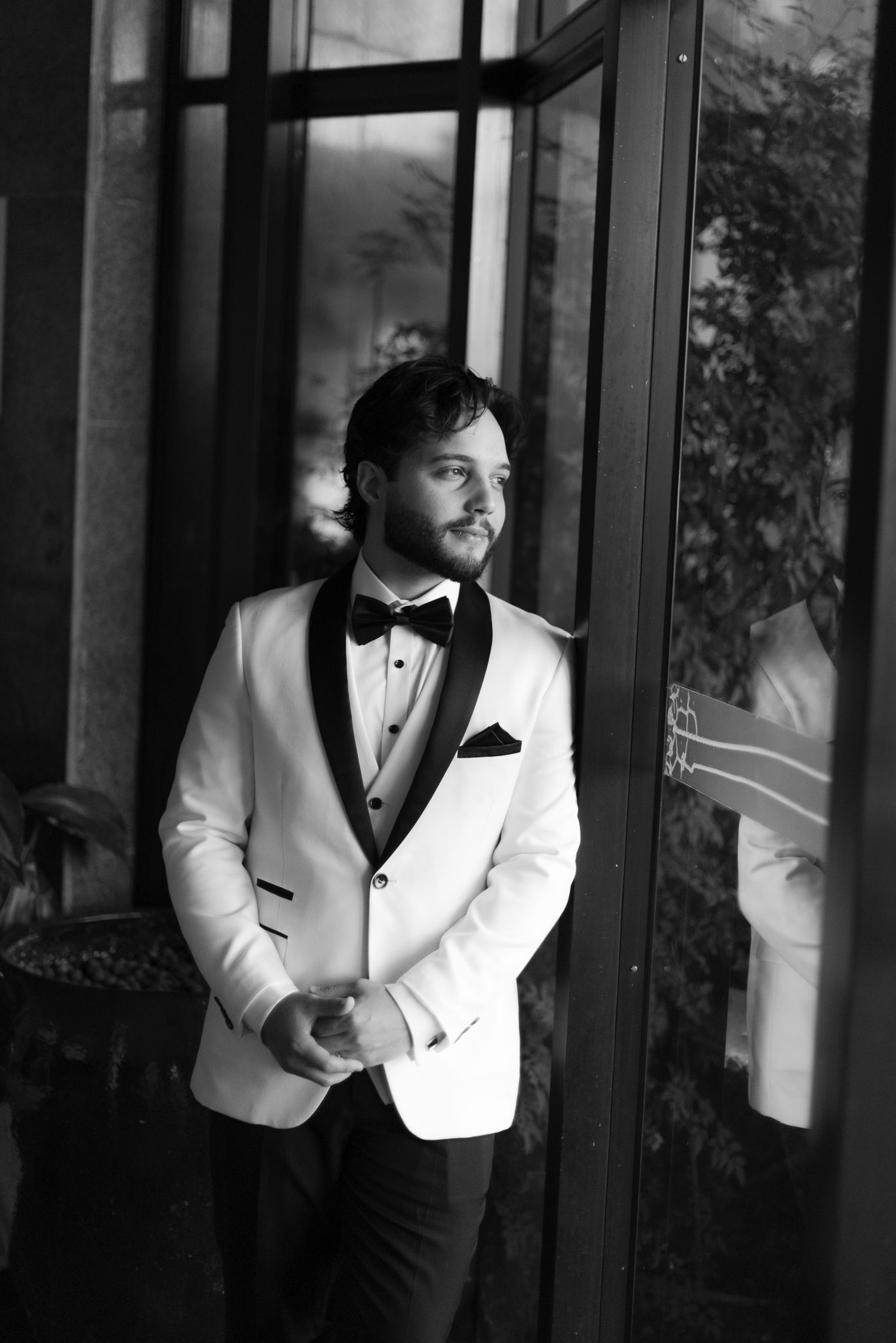 A black and white photo series showing a groom in a white tuxedo jacket and bow tie posing against a window.