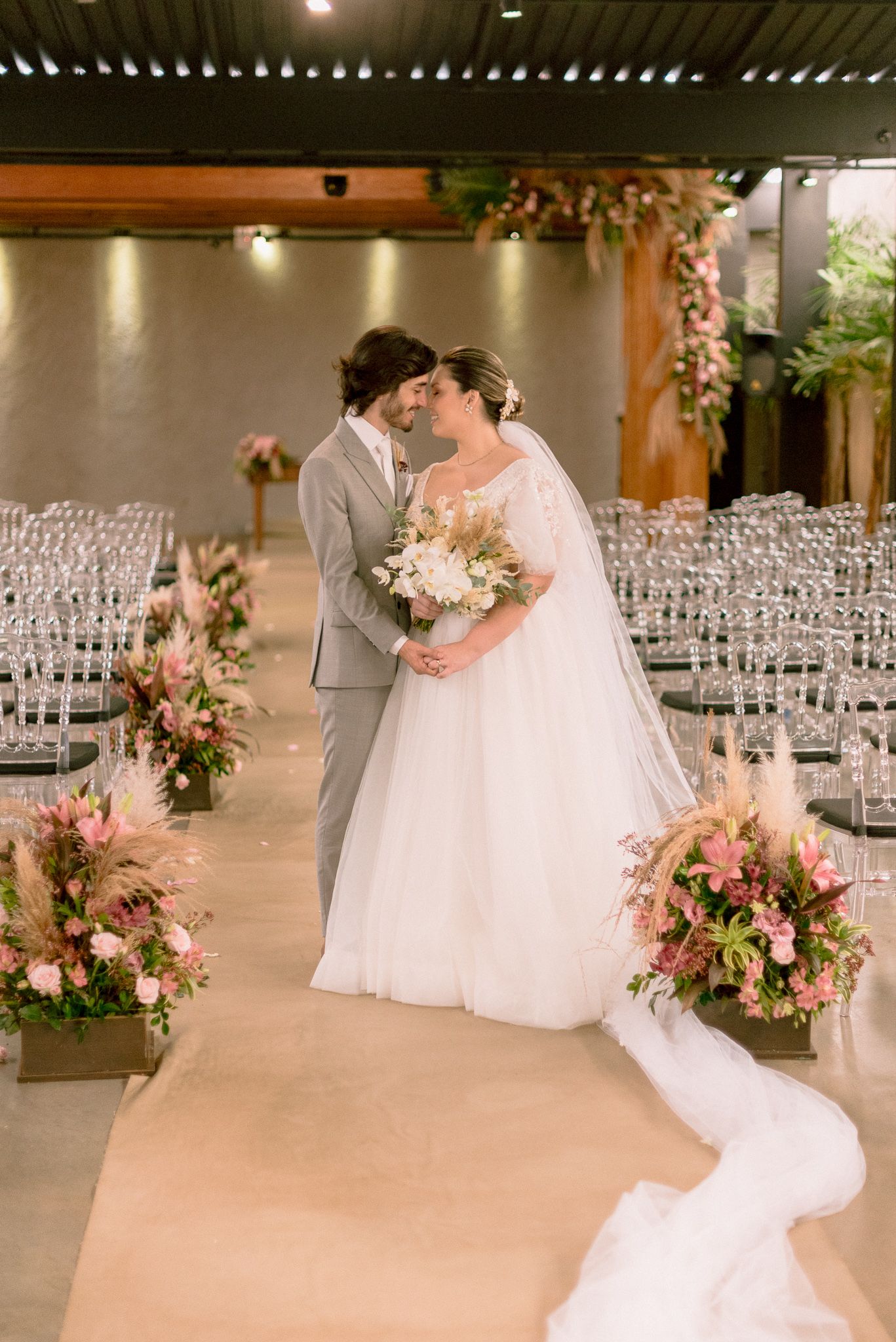 A romantic wedding couple stands in their venue among rows of white chairs and blush floral arrangements on a beige runner.