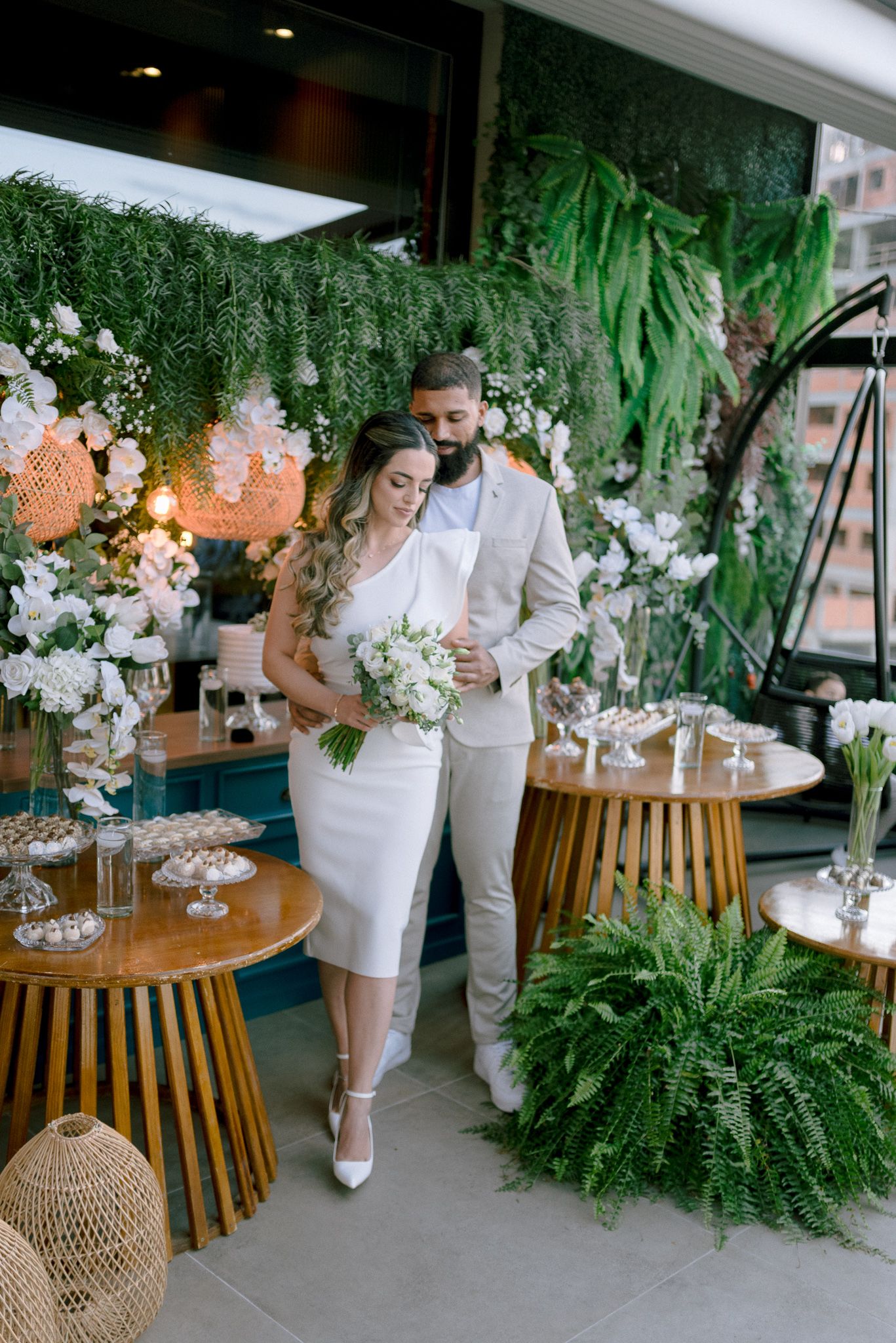 Modern wedding reception decor features white floral arrangements and lush greenery against wooden furniture and natural elements.