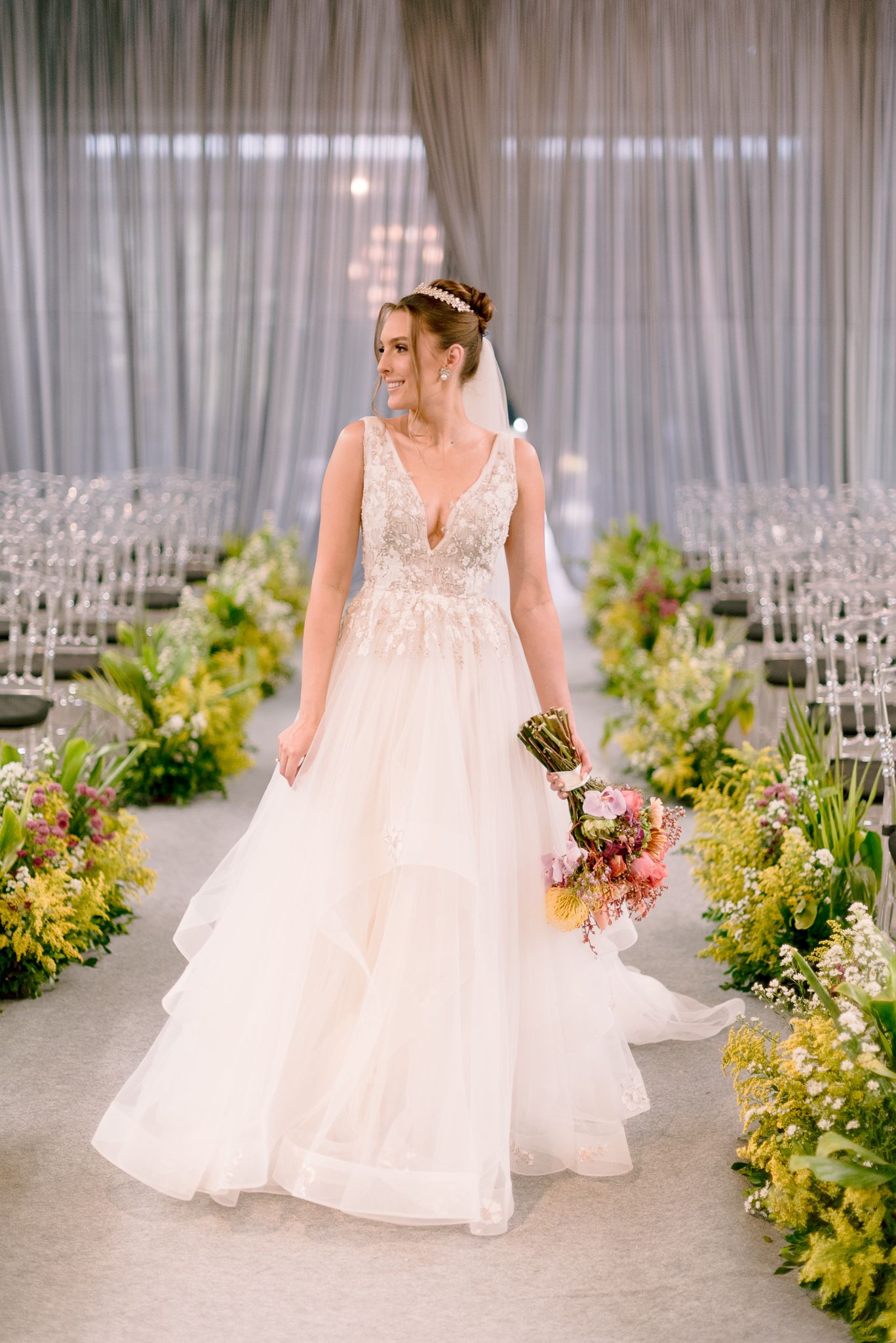 A bride in a flowing white lace wedding gown stands in a ceremony space lined with yellow and green floral arrangements.
