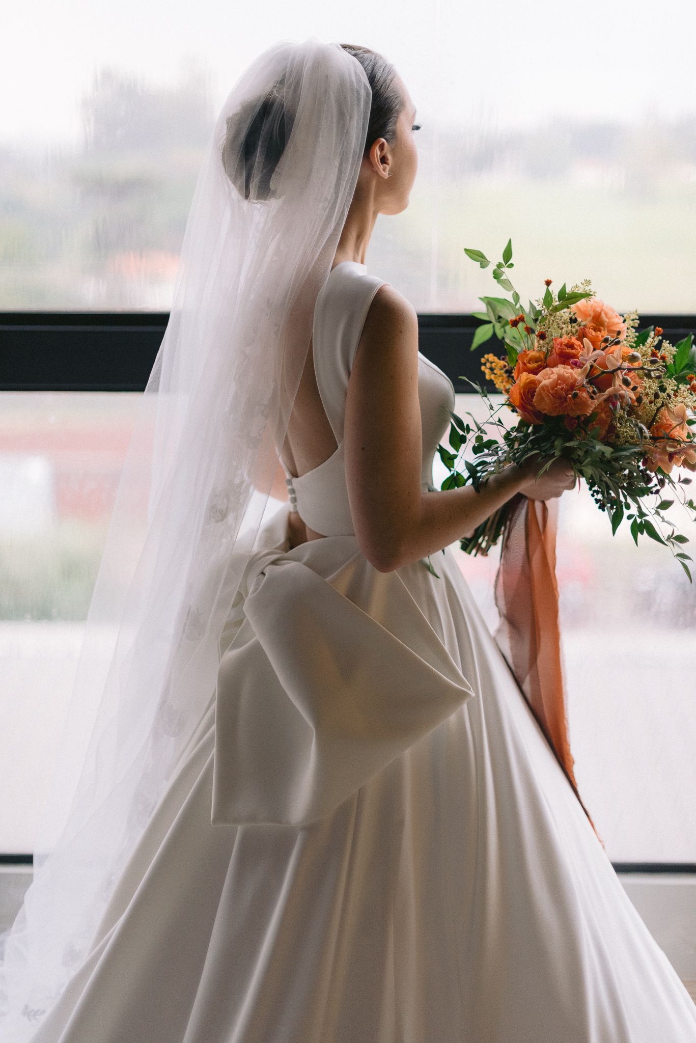 Silhouette of a bride holding a vibrant bouquet against window light in a flowing white gown.