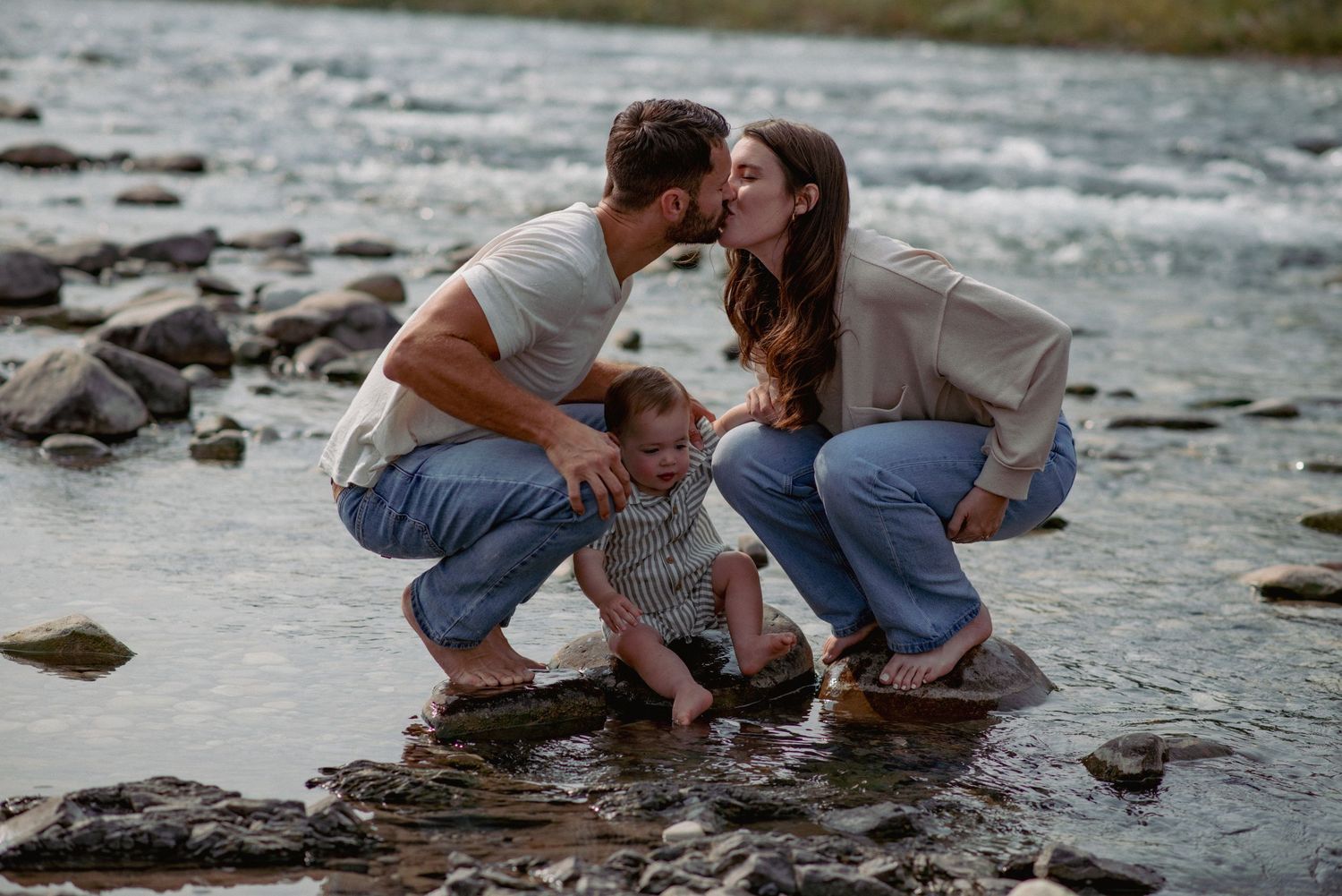 Bragg Creek family photography, parents kissing on river rocks with toddler son looking at camera